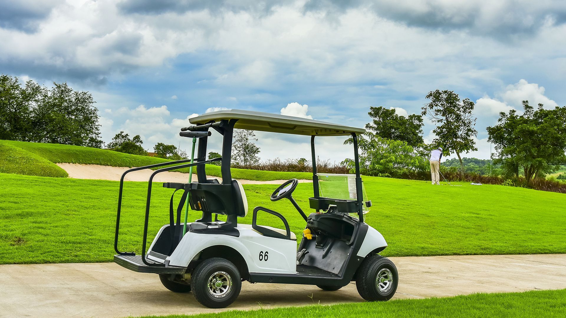 Two-seat golf cart on a paved path beside a bright green lawn and trees under a cloudy sky