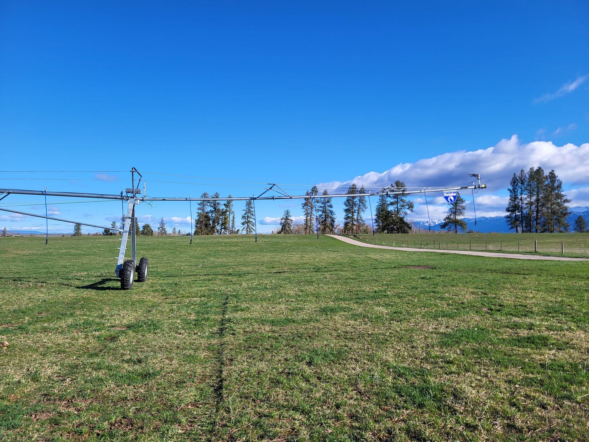 An irrigation pivot arm stretches across a sunny green field under a clear blue sky with distant trees and mountains.