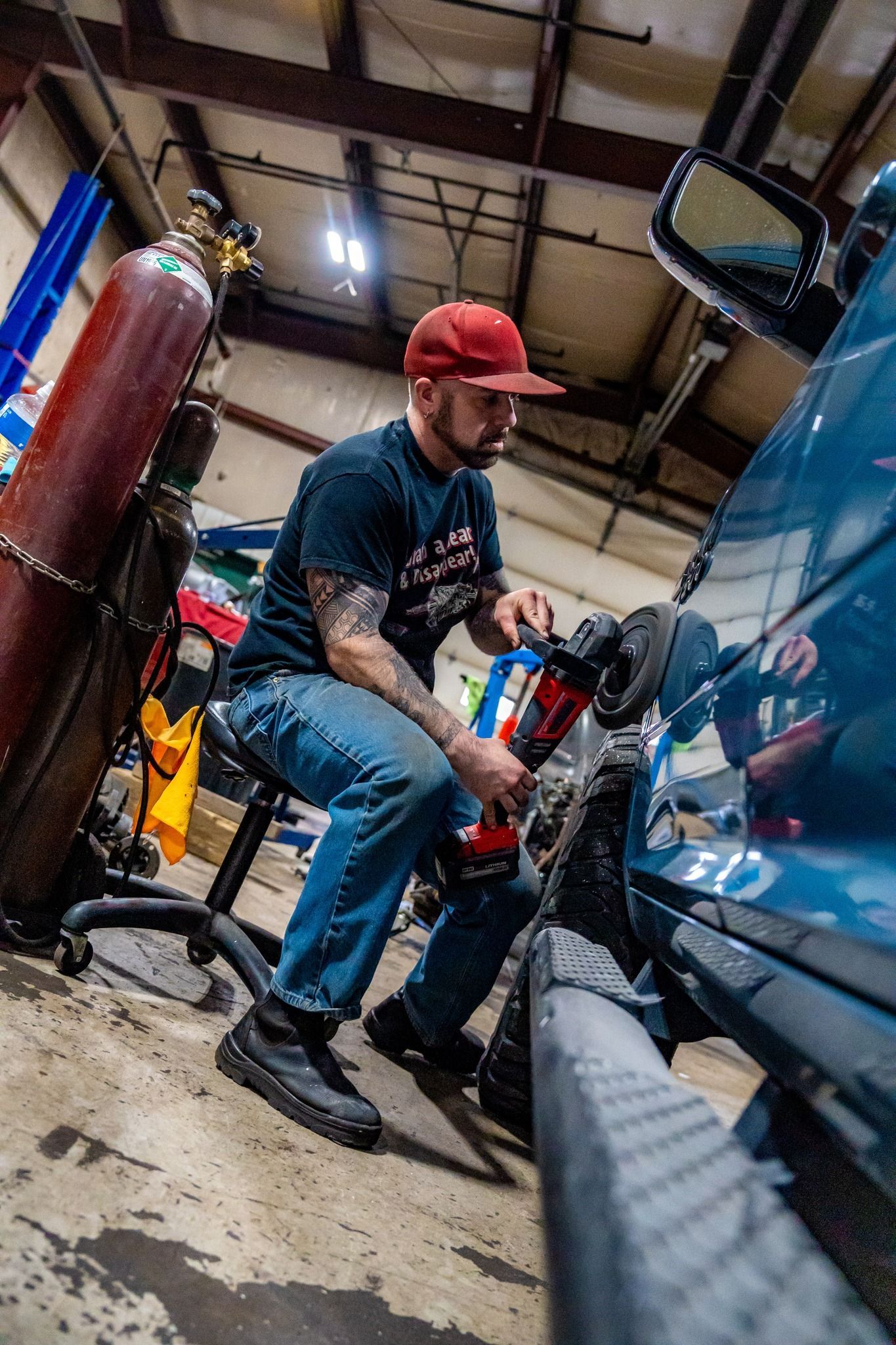 Mechanic in a red cap working on a blue car tire in a garage, with a welding tank in the background.