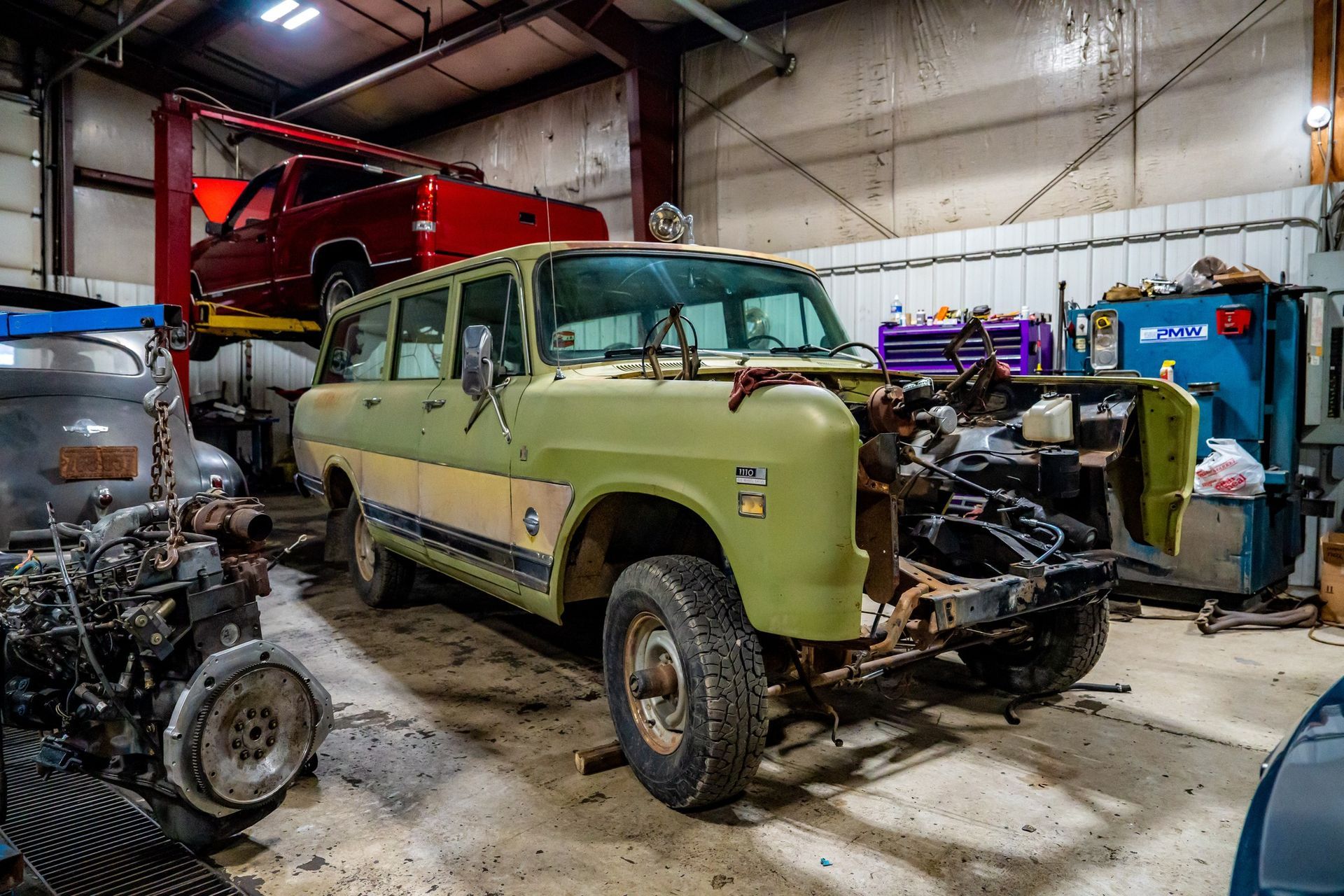 An old, green International Harvester Scout in a garage; engine work in progress, other vehicles nearby.