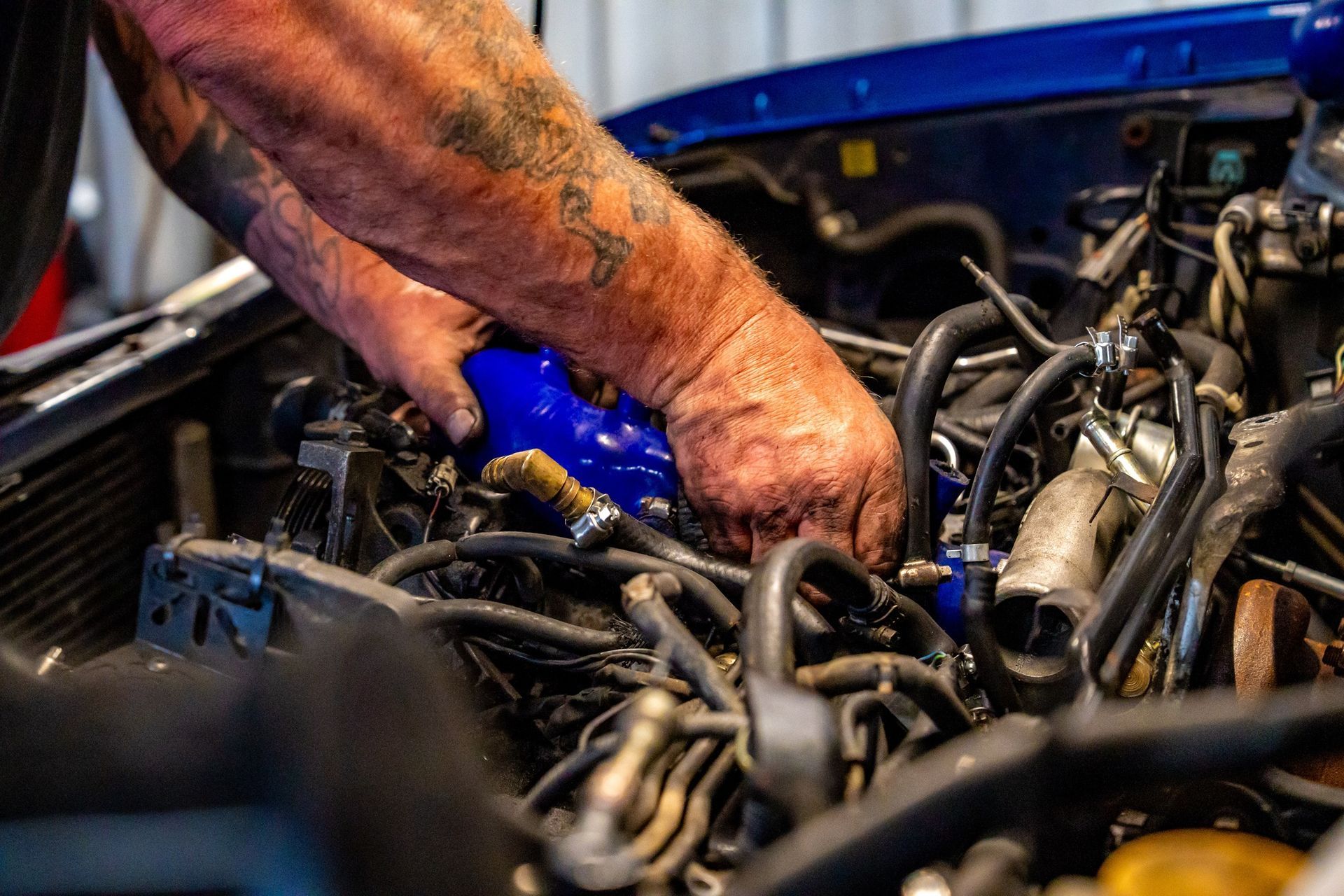 Mechanic with tattooed arms working on a blue car engine, installing a blue hose. Garage setting.