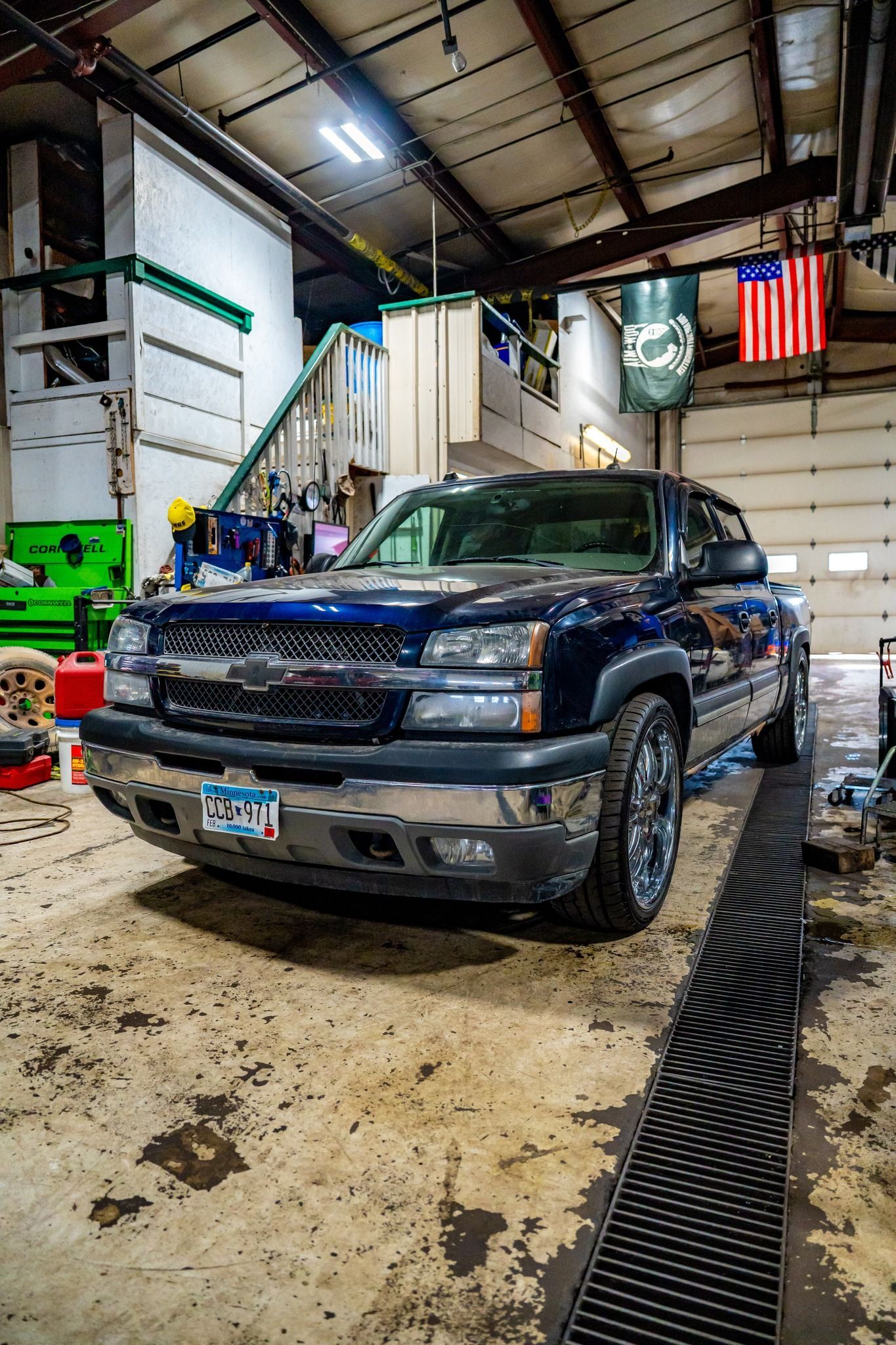 Blue Chevy Silverado pickup truck parked in a garage. American flag and a green banner hang in the background.