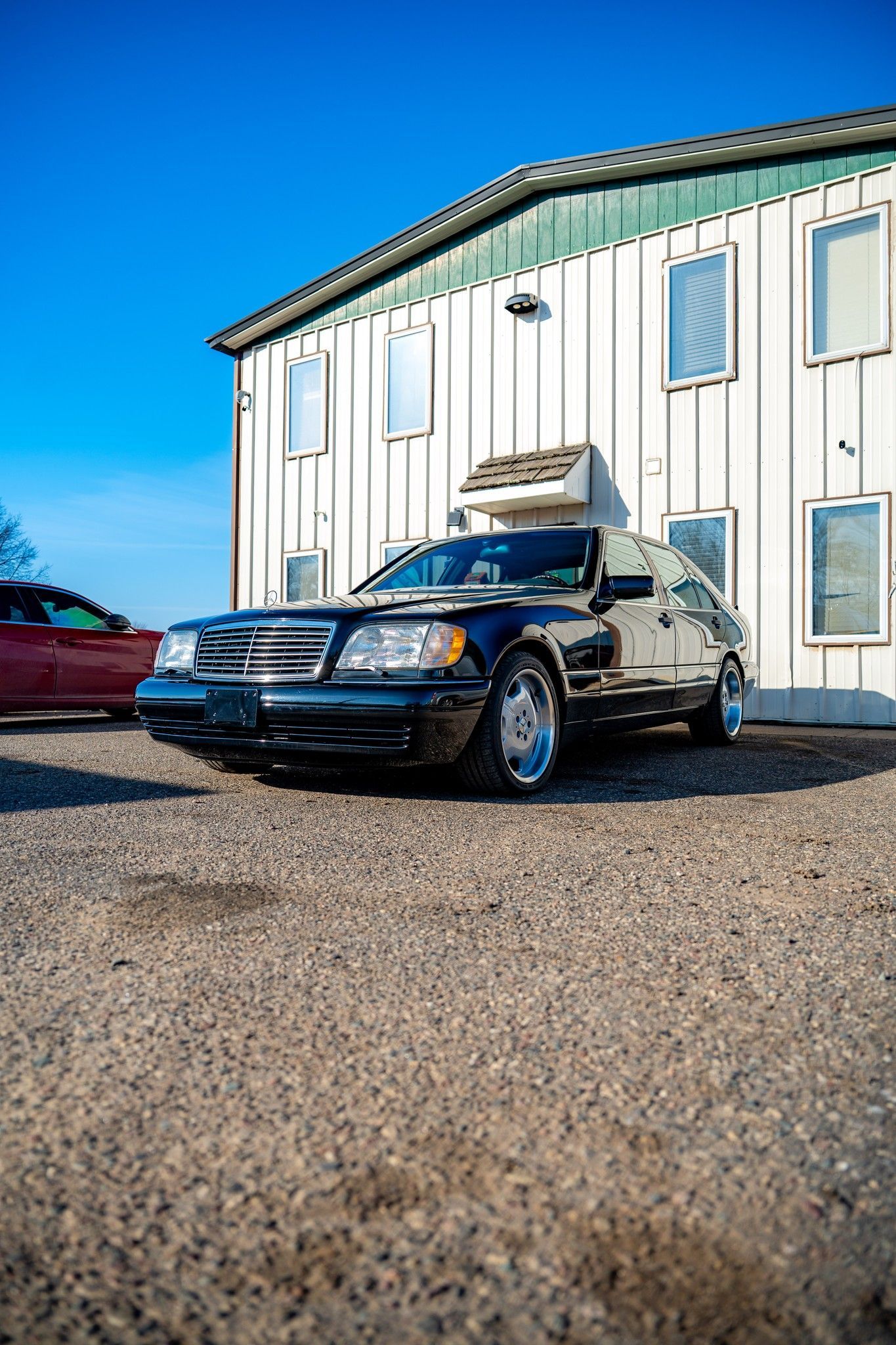 Black Mercedes sedan parked in front of a white building with blue sky. Gravel foreground.