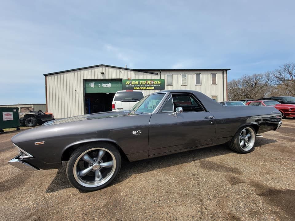 Dark gray 1960s El Camino parked in front of a building with a business sign on a sunny day.