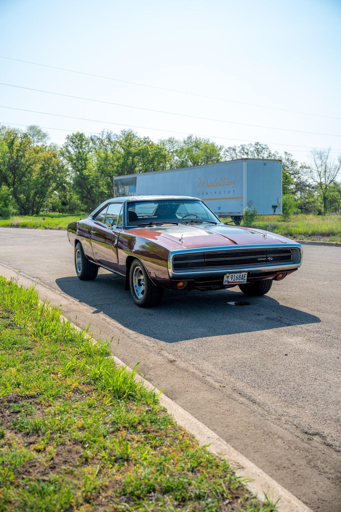 A classic red Dodge Charger parked on asphalt, in front of a white trailer, on a sunny day.