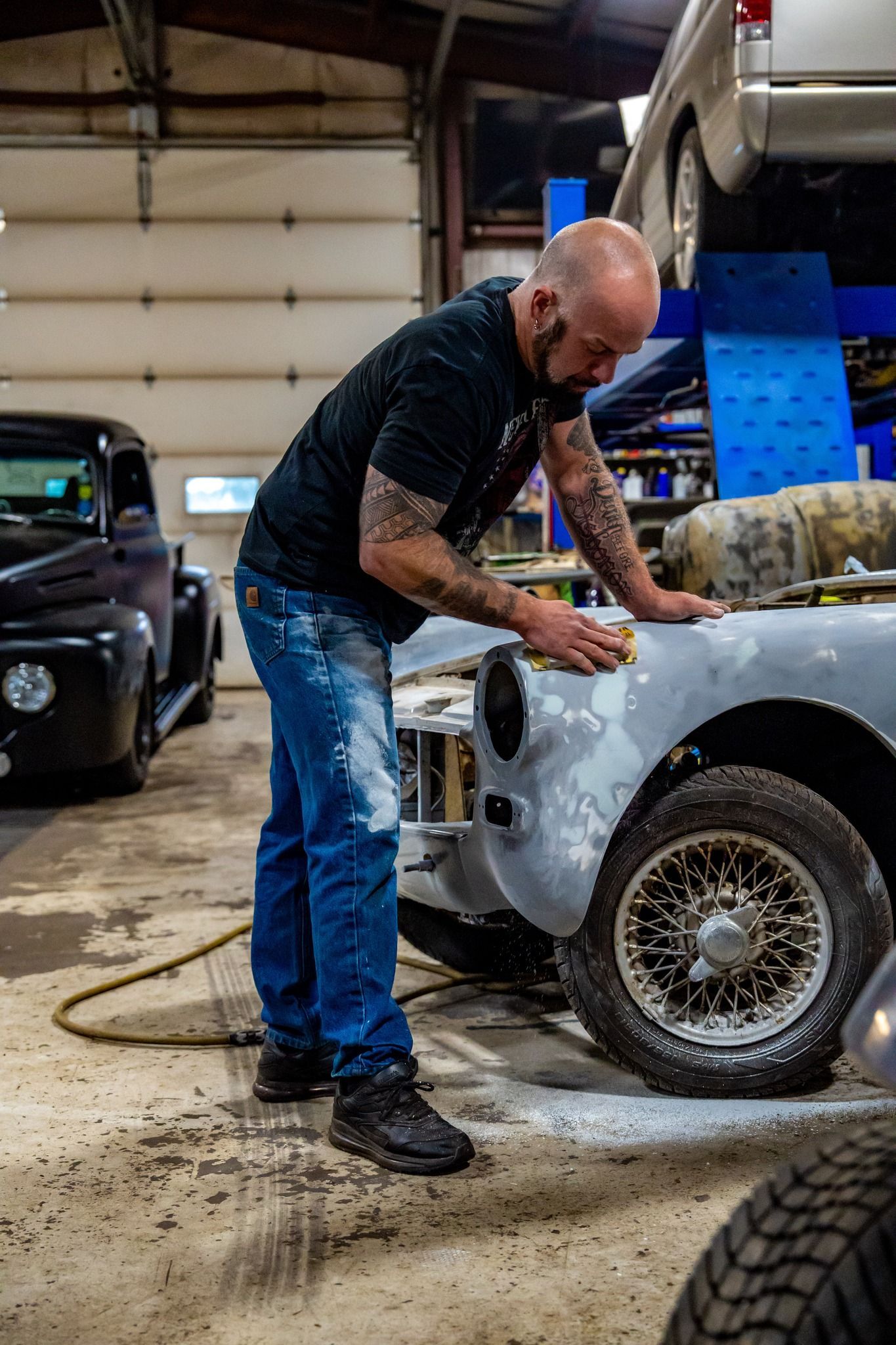 Man sanding a vintage car in a garage. He wears jeans and a black shirt, with tattoos. Another car is in the background.