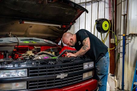 Mechanic leans over a red pickup truck engine with hood open in a garage, working on the engine.