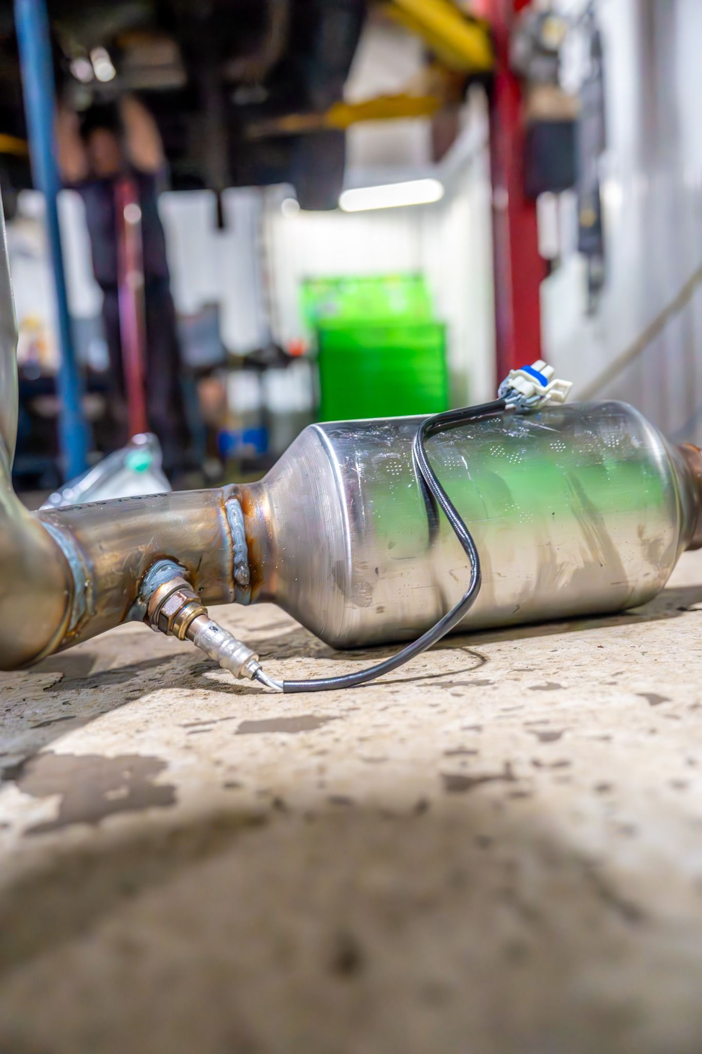 Catalytic converter on a garage floor. It is silver with a green tint, attached to a metal pipe with a sensor.