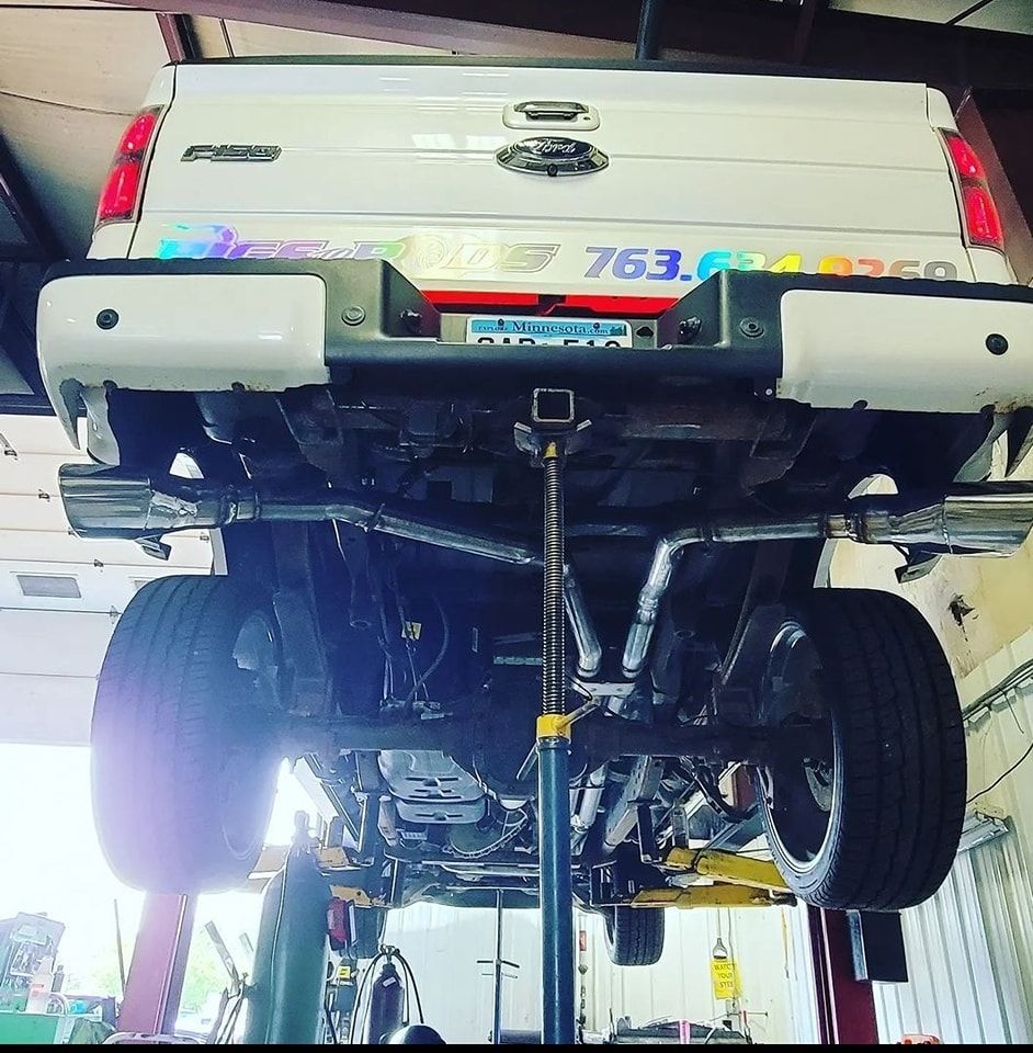 White truck on a lift with custom exhaust. Underside view showing the vehicle's mechanics and a tow hitch.