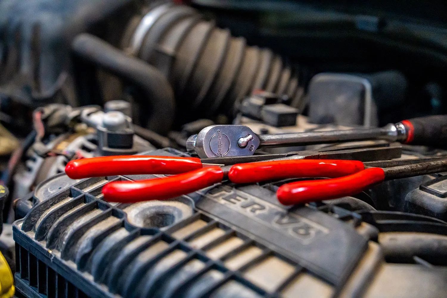 Red pliers and a wrench sit on a car engine, implying repair or maintenance.