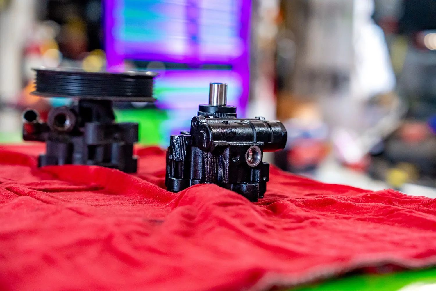 Two black power steering pumps on a red cloth, with a blurred shop background.
