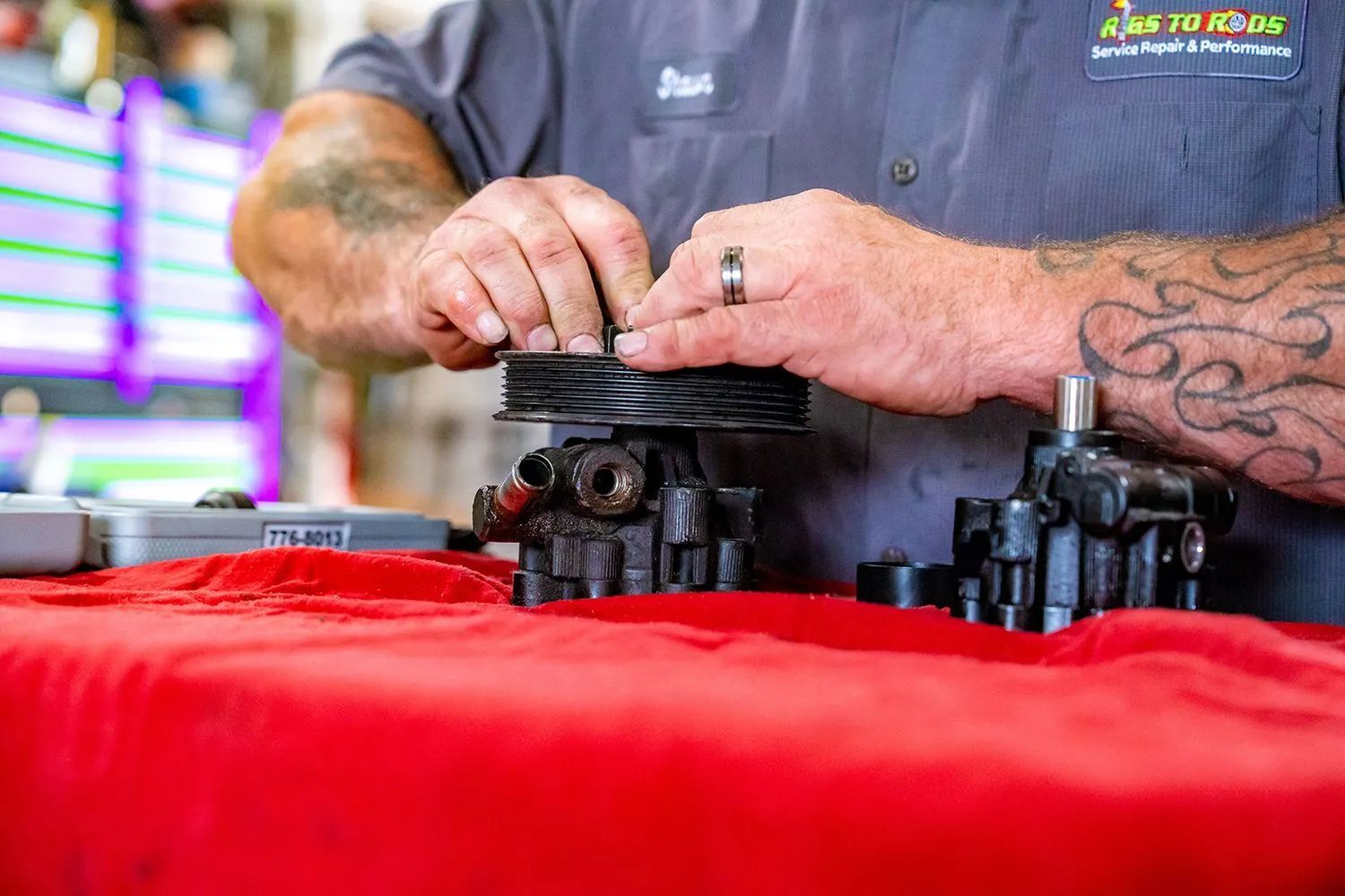 Mechanic assembling a car part on a red surface; tattoos visible.