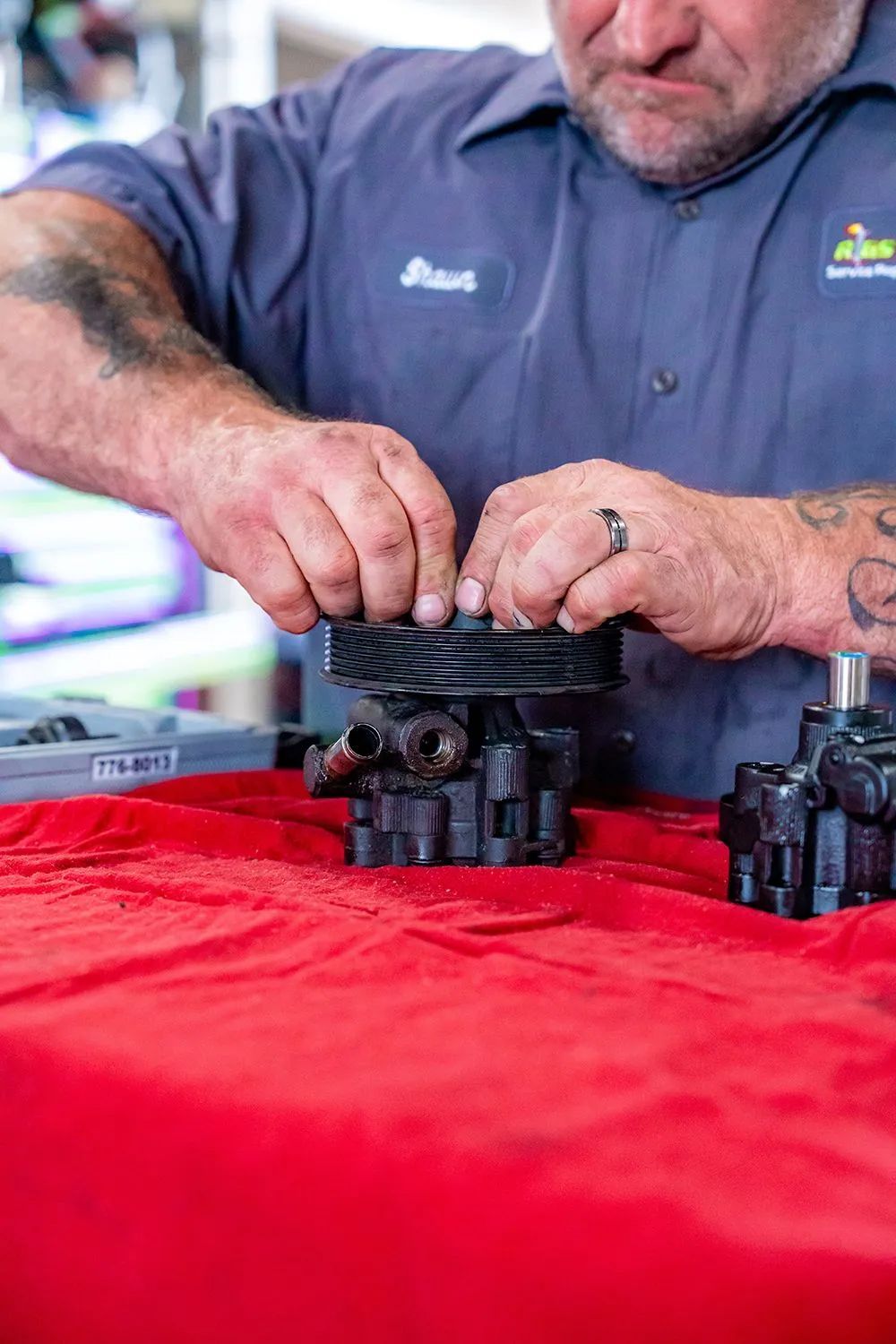 Mechanic in blue shirt works on a black mechanical part on a red surface.
