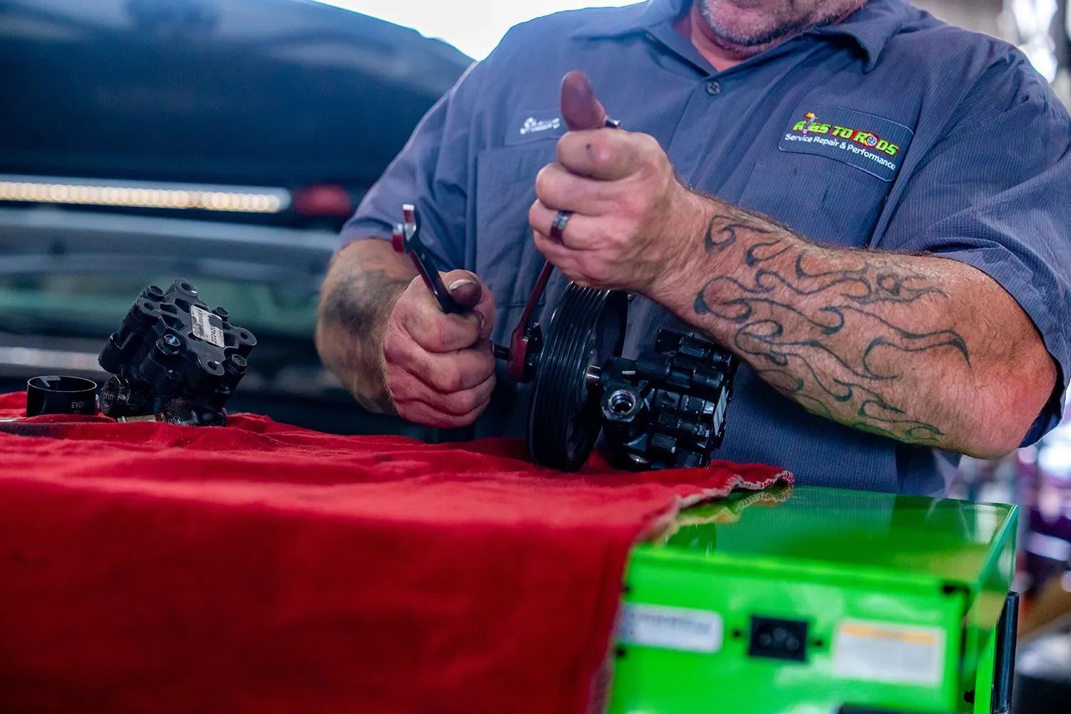 Mechanic with tattoos working on a car part at a workbench with green equipment.