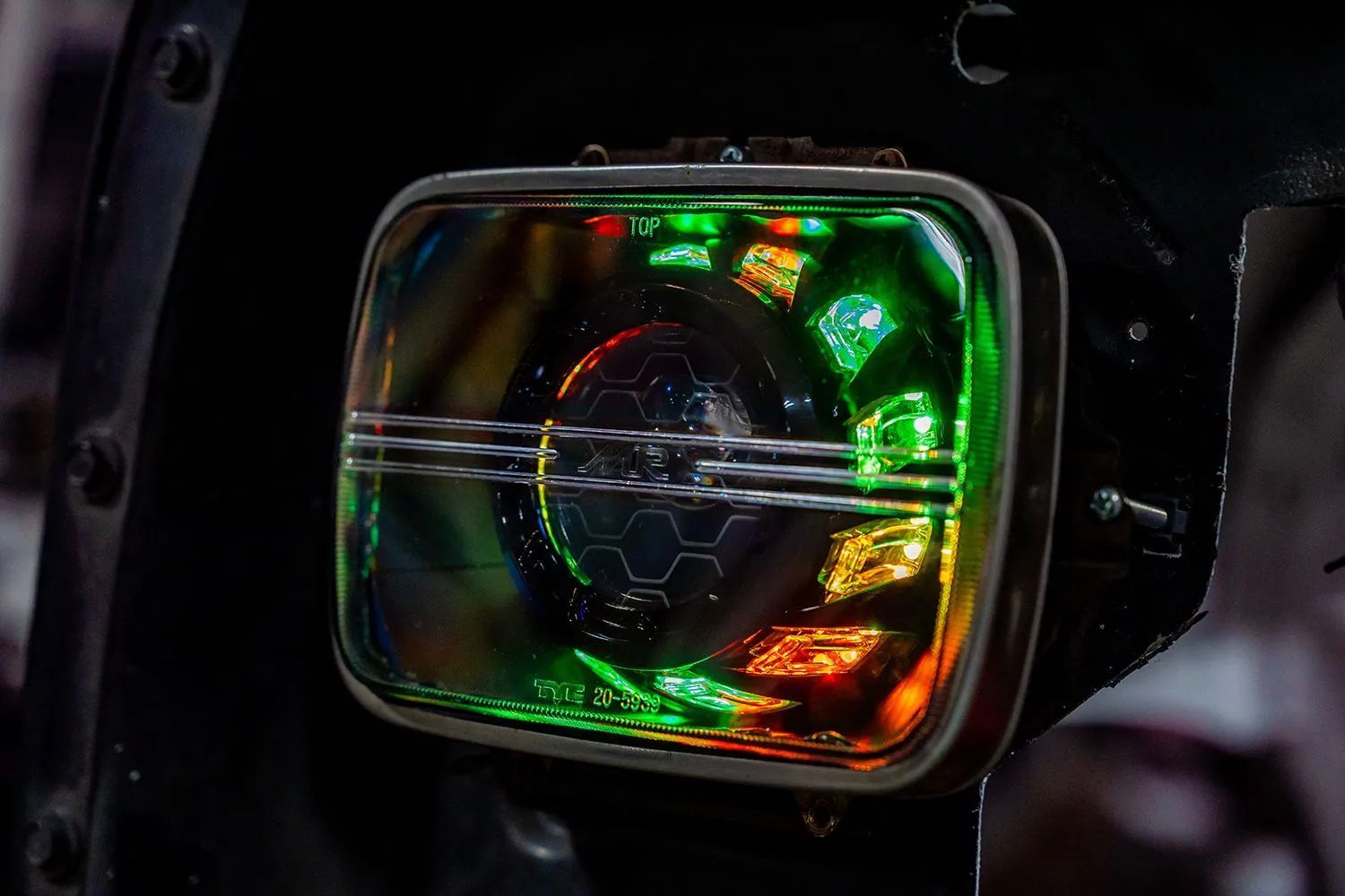 Square headlight with green, orange, and yellow LED lights and a chrome trim on a dark background.