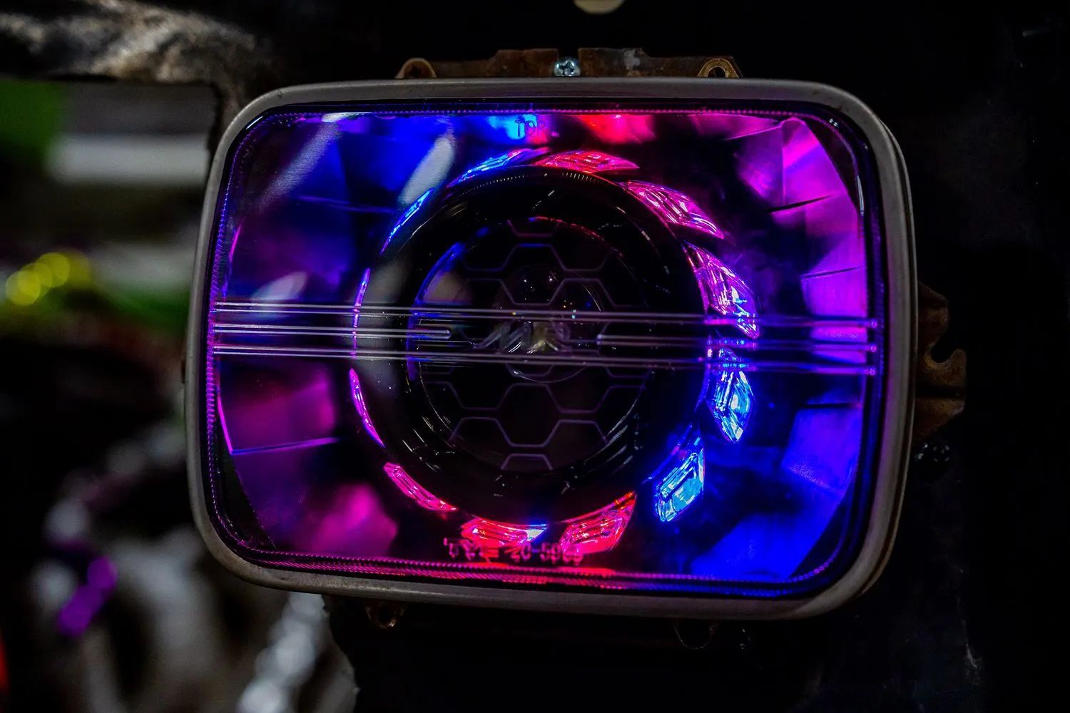 Close-up of a rectangular LED headlight with a hexagonal pattern, glowing in vibrant pink and blue.