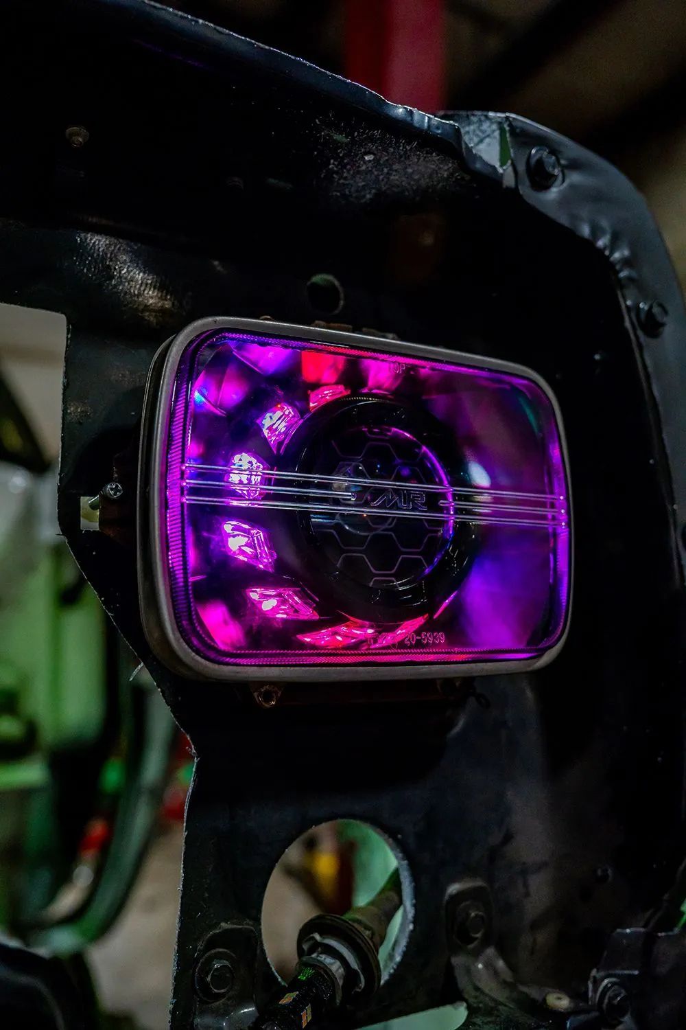 Rectangular car headlight with a purple and pink glowing interior, mounted on a black vehicle frame.