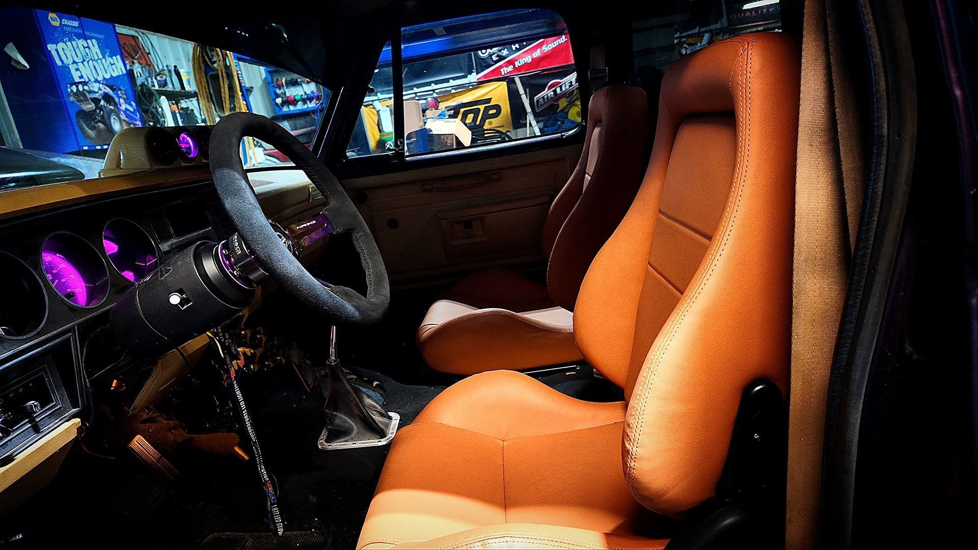 Interior of a vintage car with brown and white bucket seats, purple dashboard lights, and a black steering wheel.