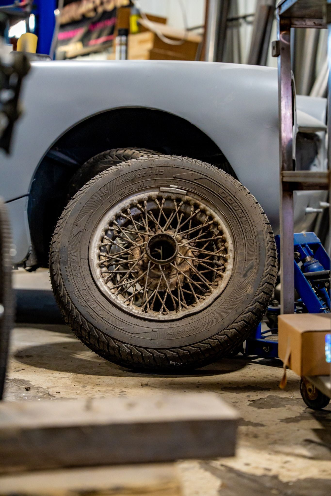 A vintage car tire with wire spokes sits in a workshop next to the car's body.