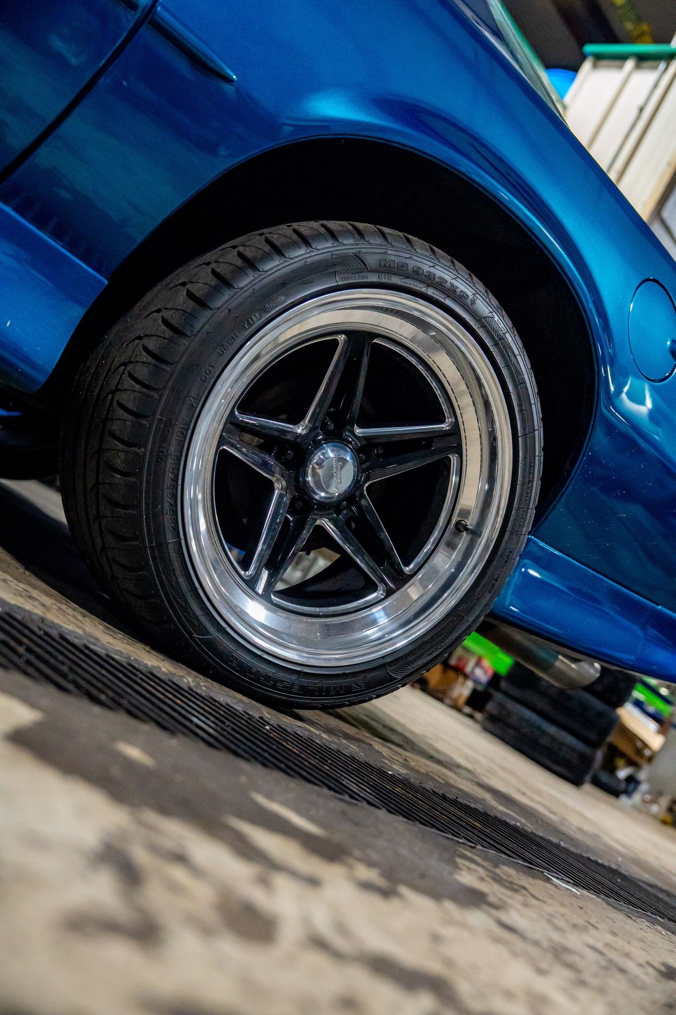 Blue car wheel with black tire and star-shaped silver and black rim on a gray surface.