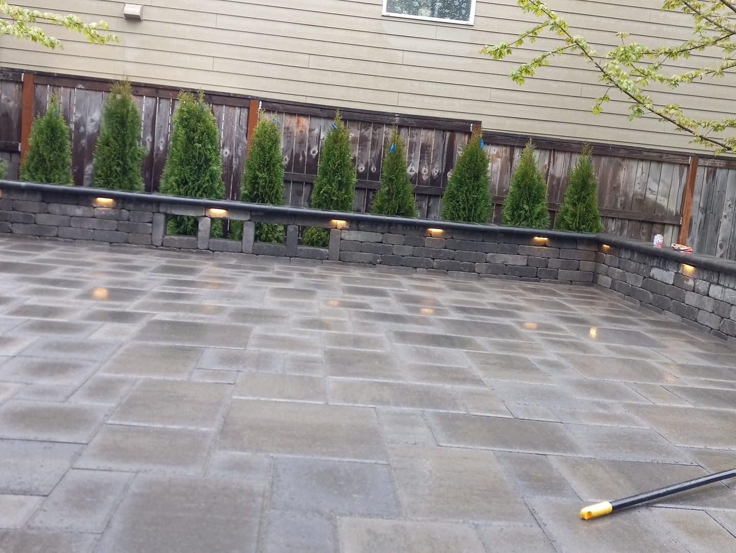 Stone patio with retaining wall, evergreen trees, and recessed lighting, against a wooden fence.