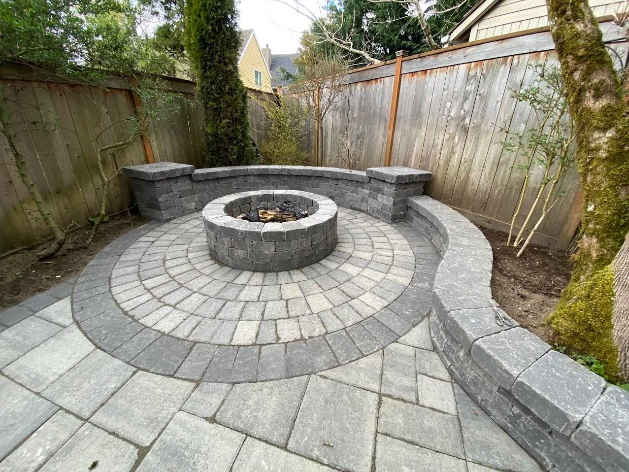 Circular paved patio with a fire pit, surrounded by a low wall and wooden fence.