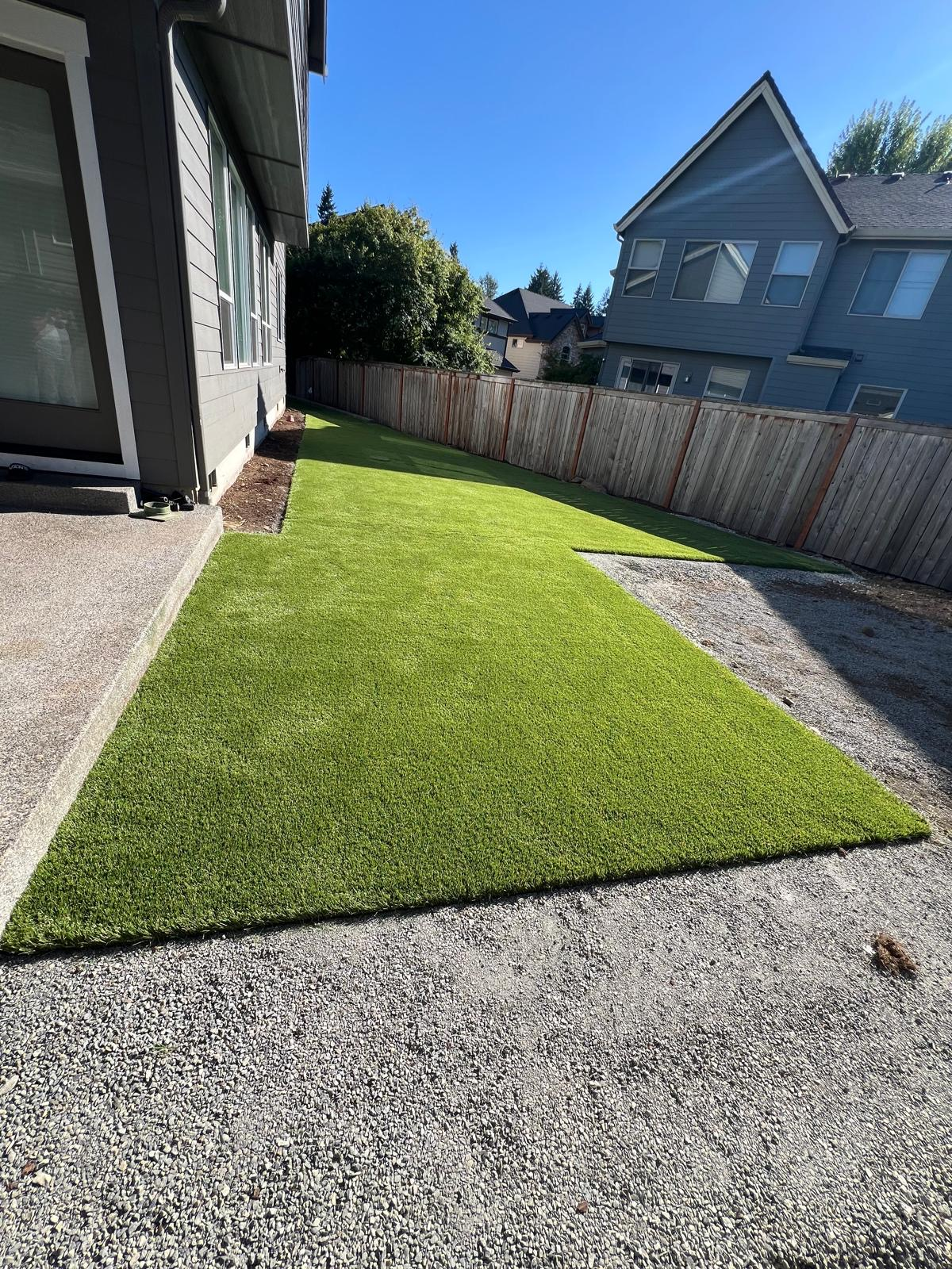 Backyard with green artificial turf, gravel, a fence, and a gray house on a sunny day.