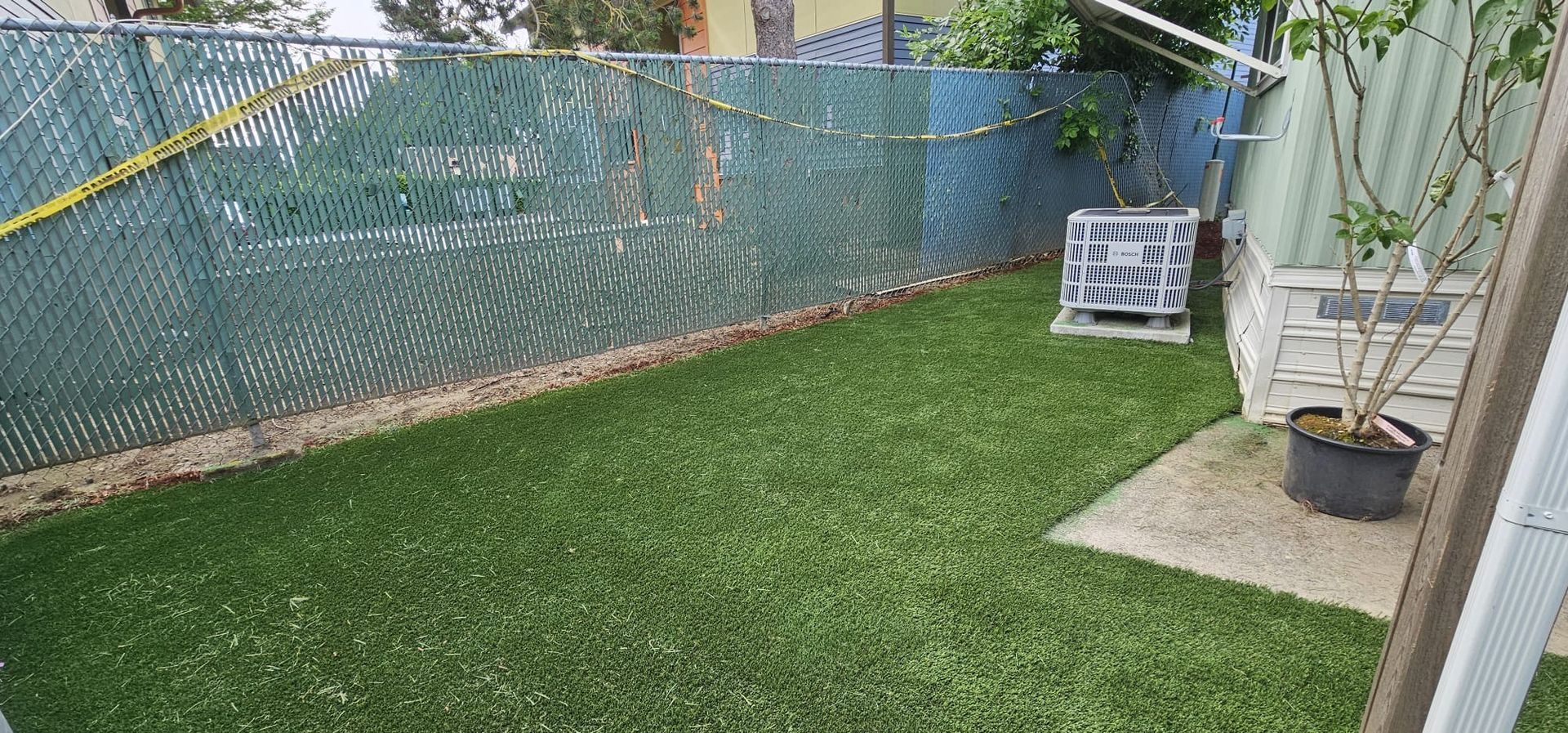 A backyard with green artificial turf, a green chain link fence, and a potted plant.