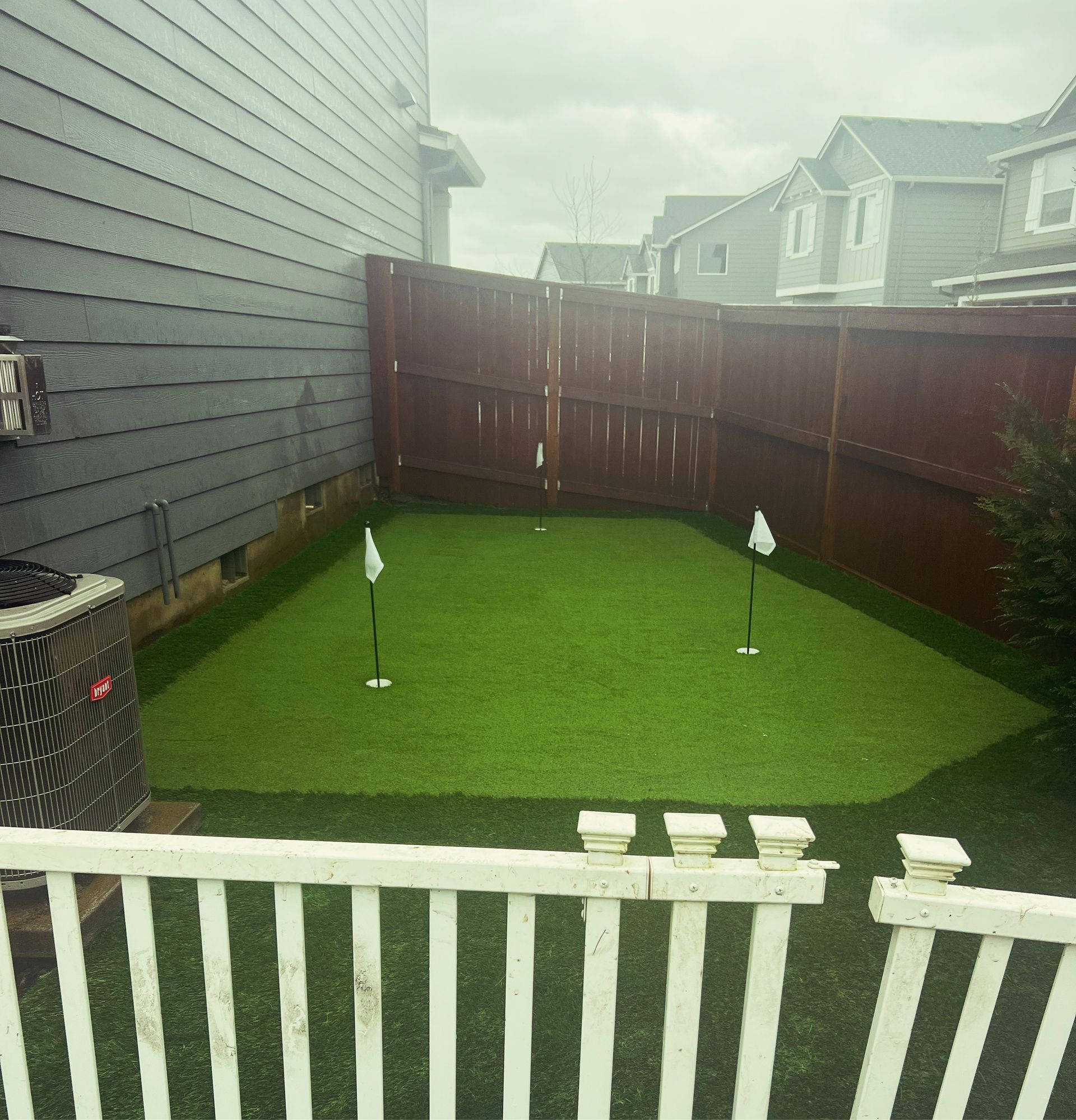 Small backyard with artificial turf and two golf holes. Fenced in with houses in the background.
