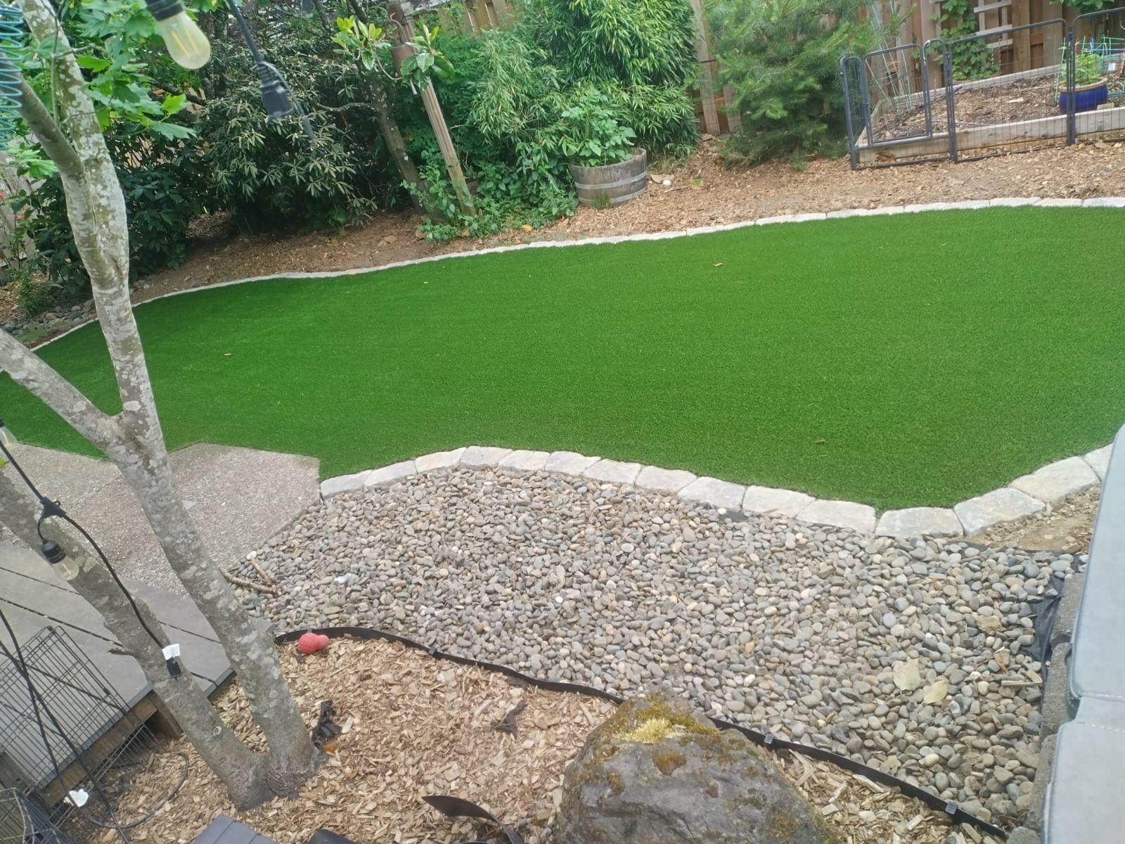Artificial grass lawn bordered by gravel and garden bed, surrounded by trees and greenery.