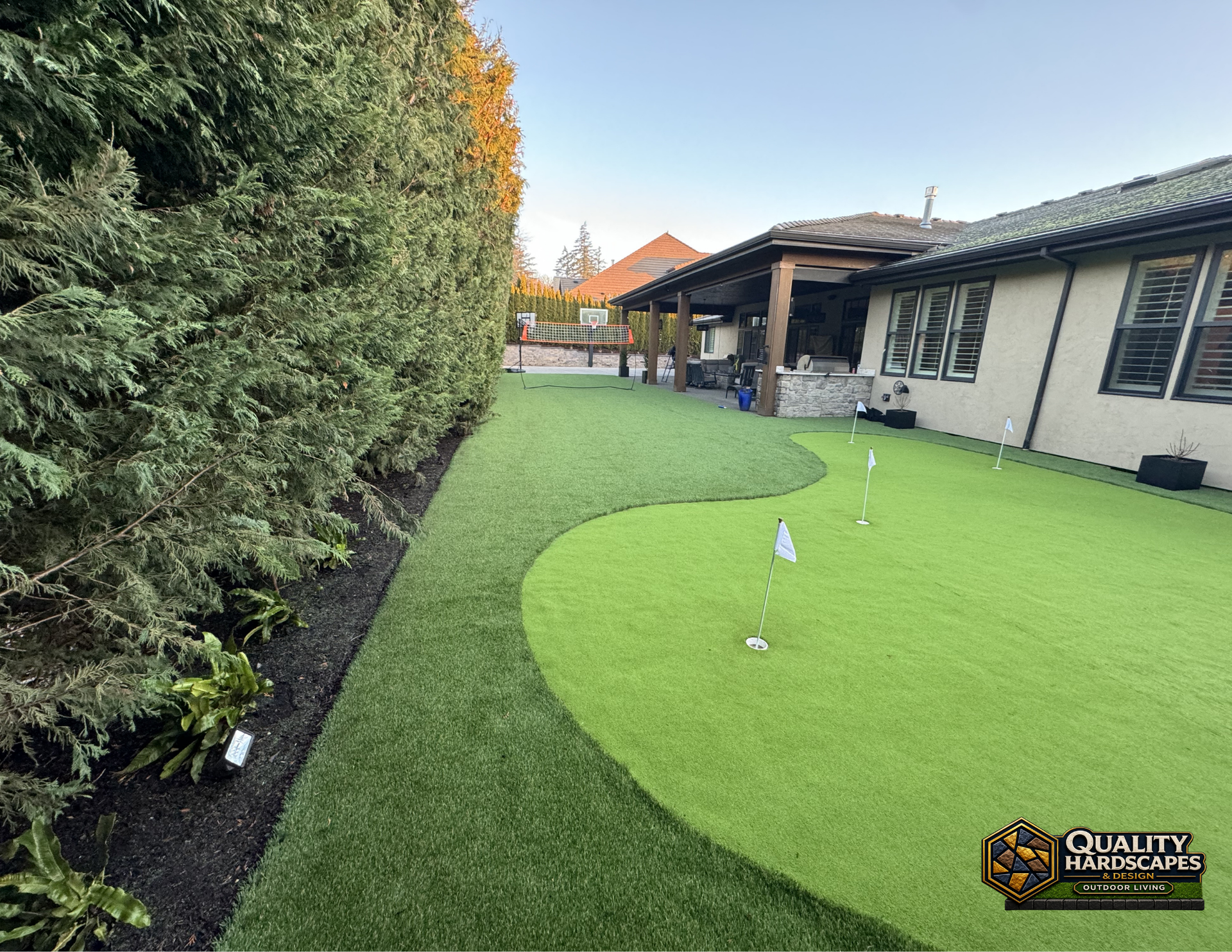 Neatly landscaped backyard with artificial turf lawn, tall hedge, and covered patio beside a house