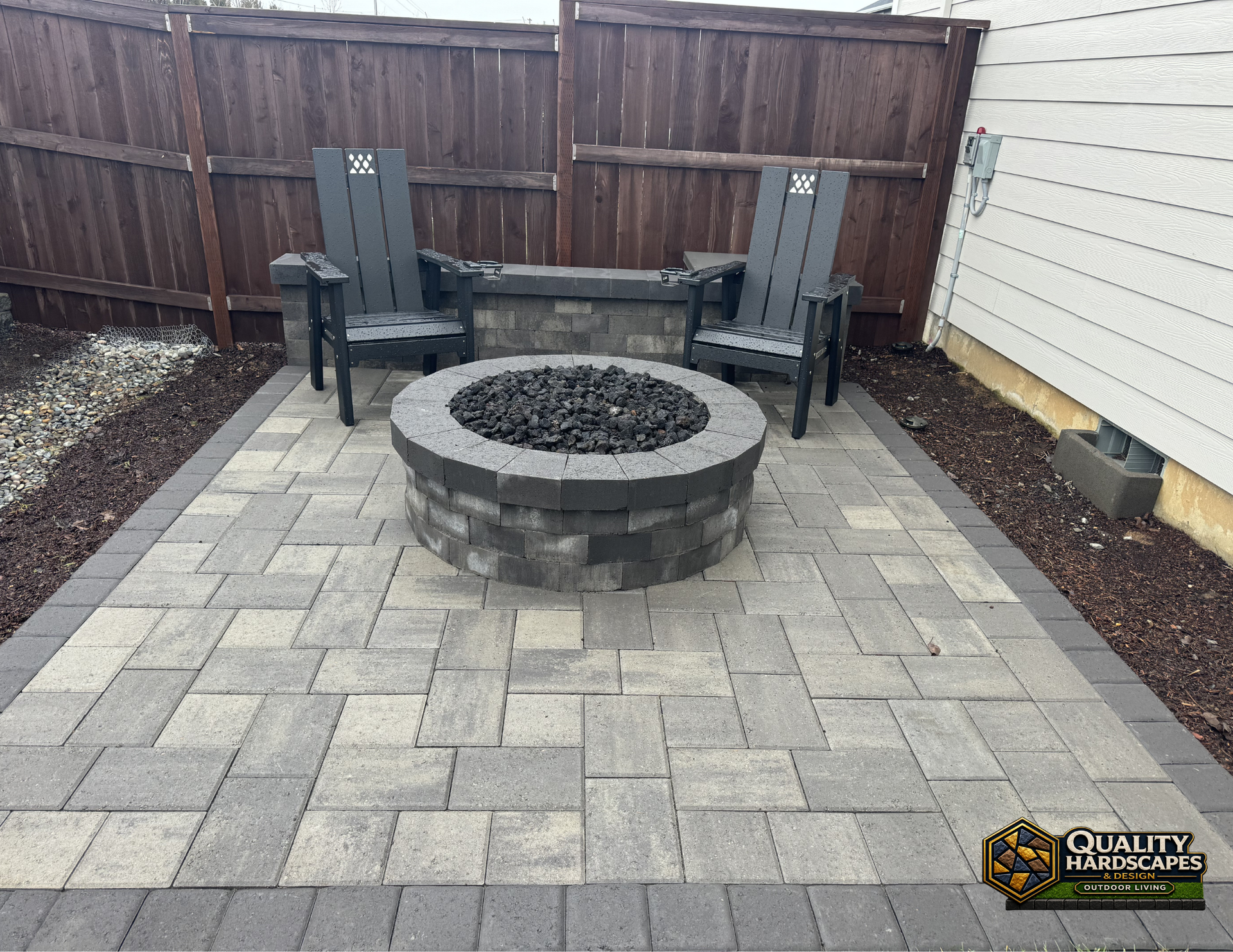 Backyard patio with paver stones, a circular fire pit, and two chairs against a wooden fence.
