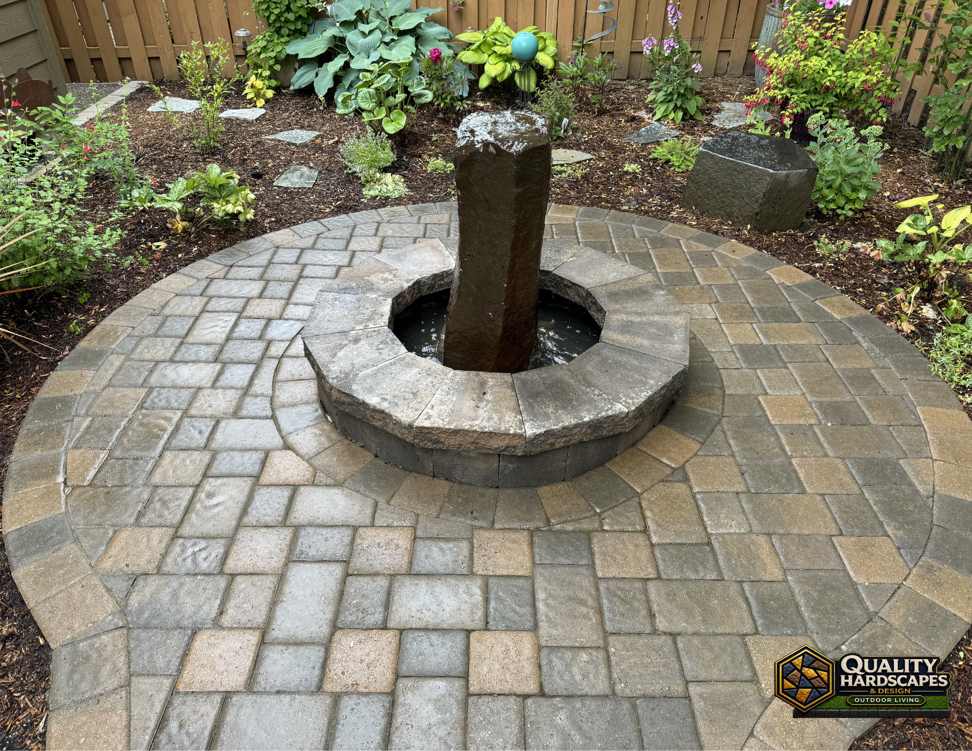 Circular stone fountain in a paved garden courtyard with surrounding plants and flowers