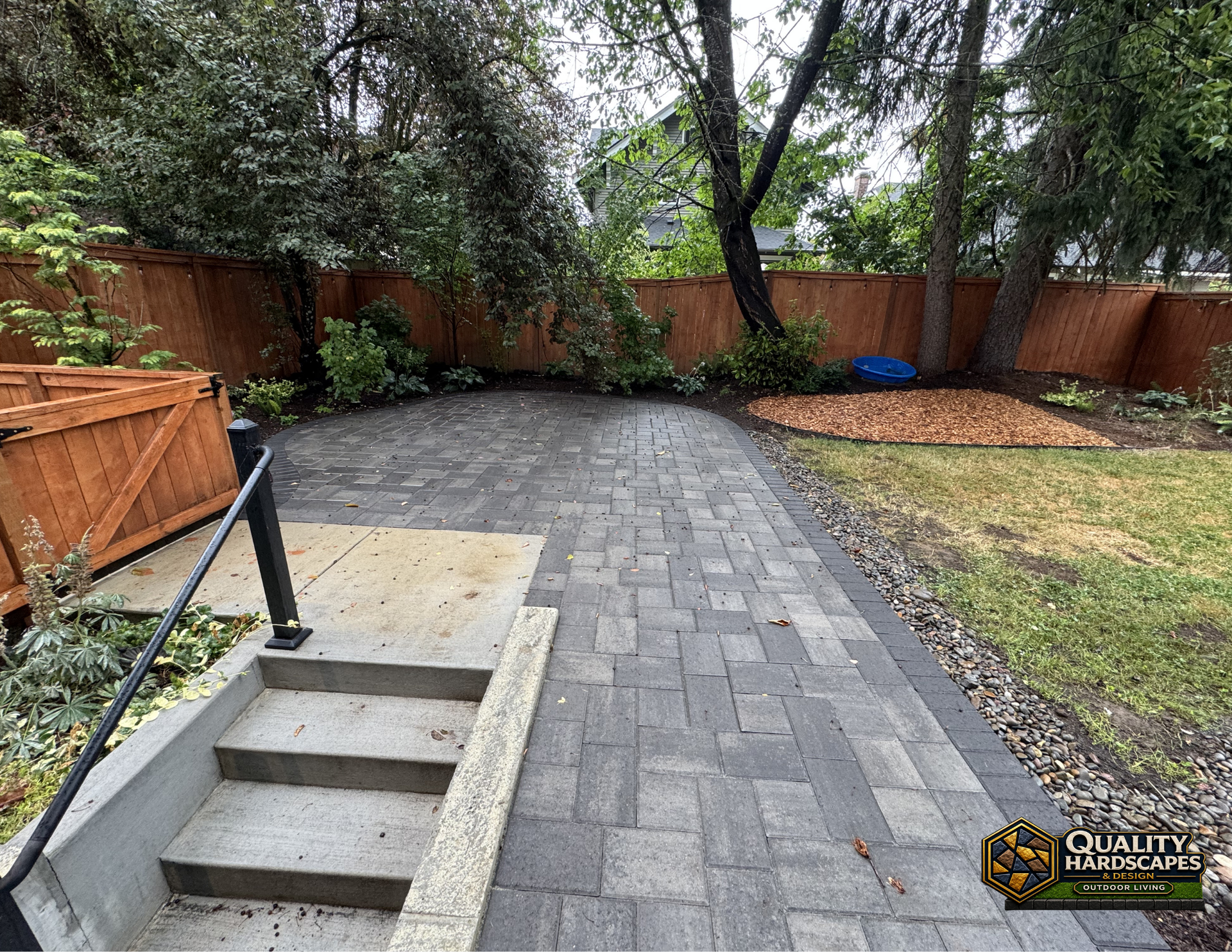 Backyard with gray paver patio, brick fence, trees, and a small concrete stairwell by a wooden deck