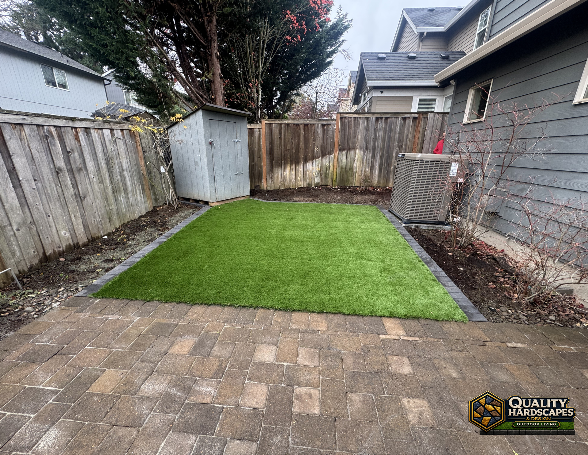 Small backyard with a green lawn, stone patio, wooden fence, and gray shed beside a house.