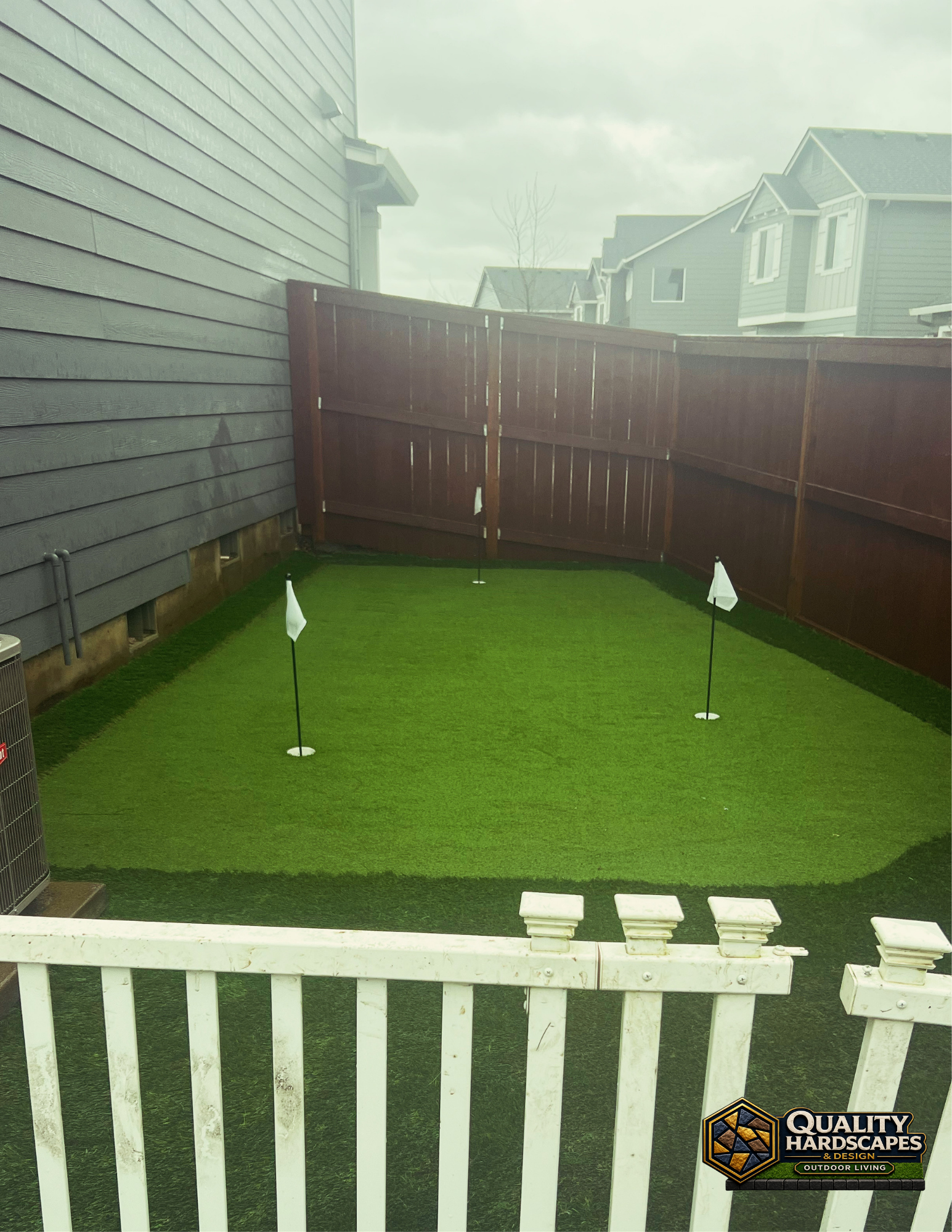 Small fenced backyard putting green with two flagsticks beside a white deck railing