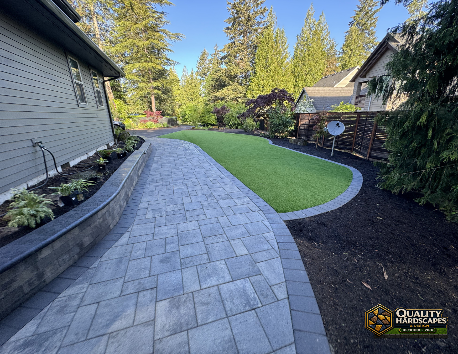Curved gray paver walkway beside a house, edging a green lawn and landscaped yard