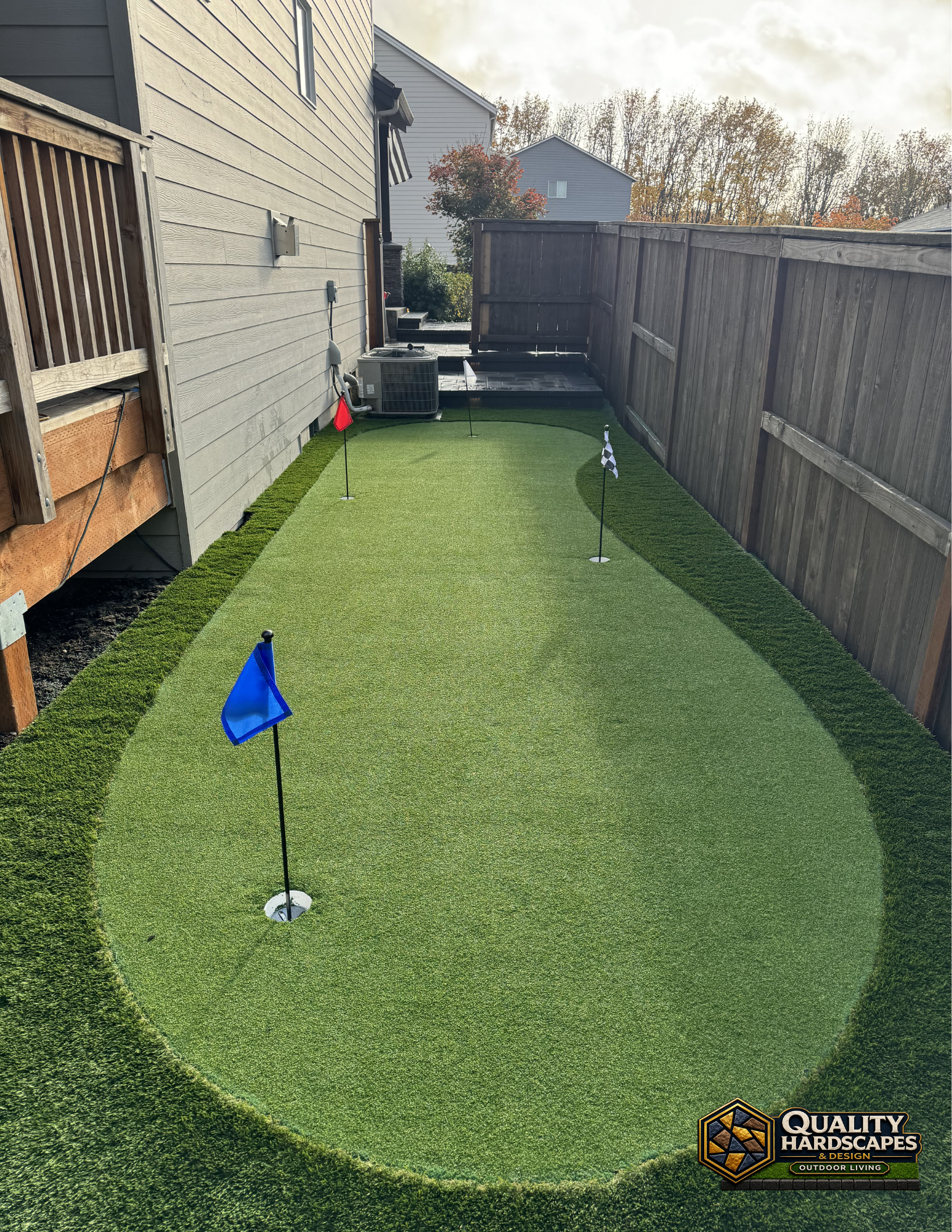 Narrow backyard putting green beside a house and fence, with a blue flag and a golf ball near the cup