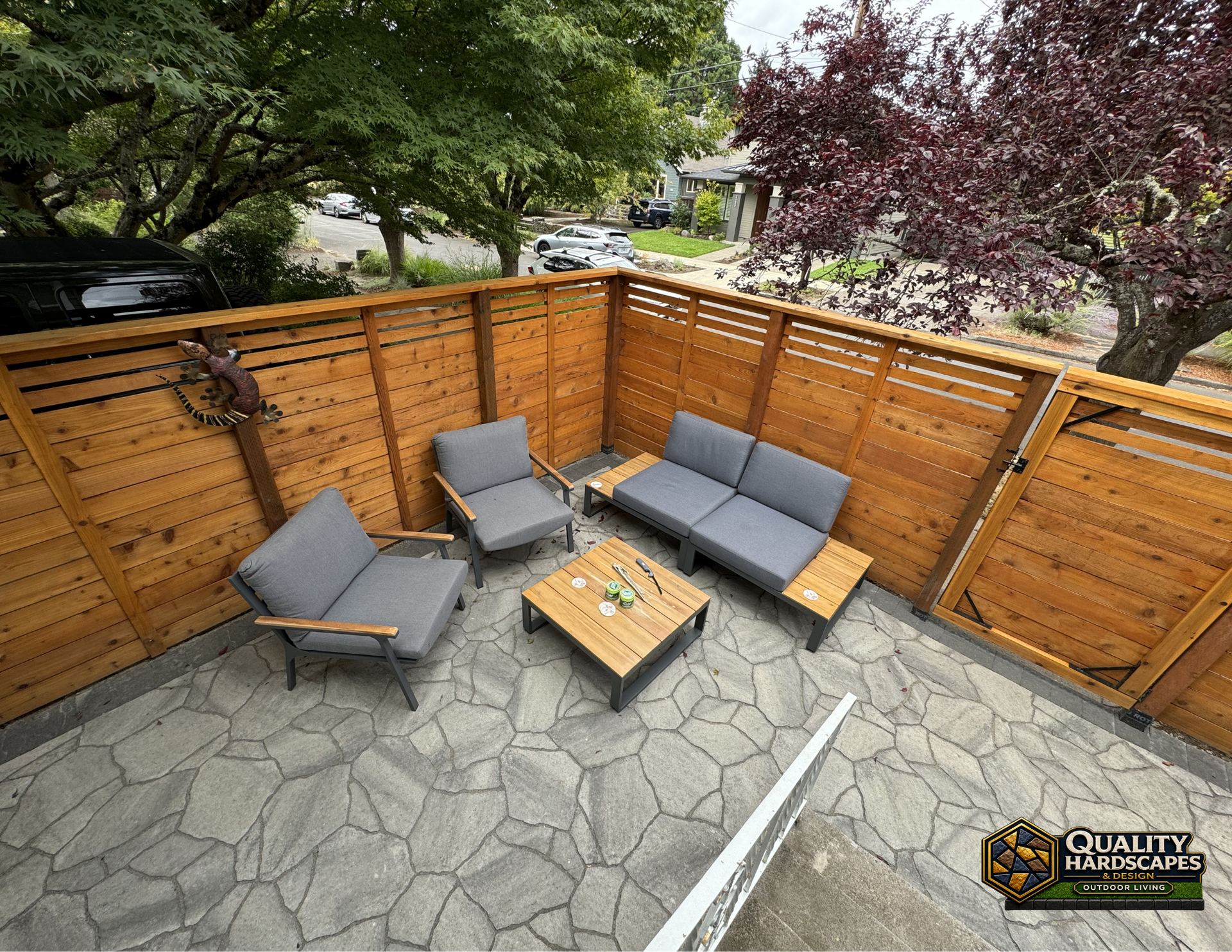Small patio with gray outdoor seating, wooden fire table, and privacy fence surrounded by trees.