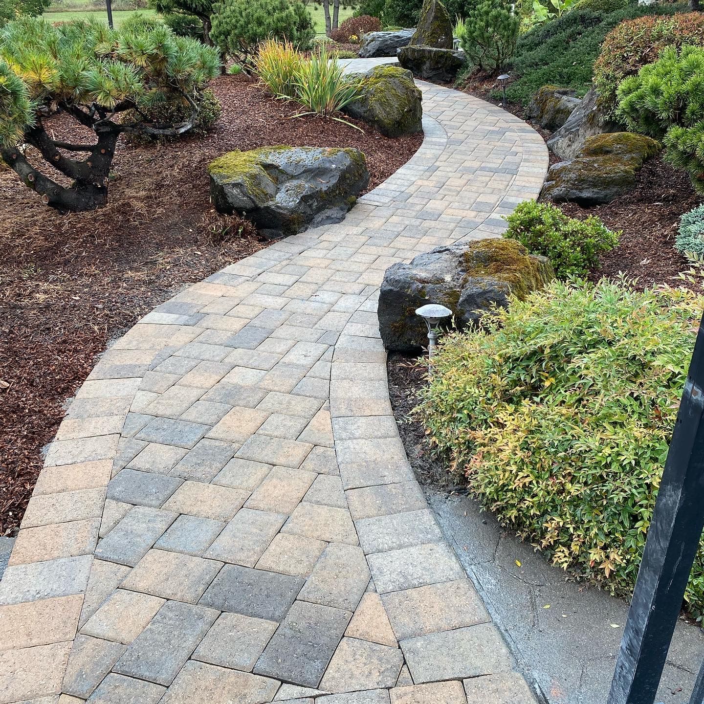 Stone paved path winds through a garden with rocks, shrubs, and mulch.