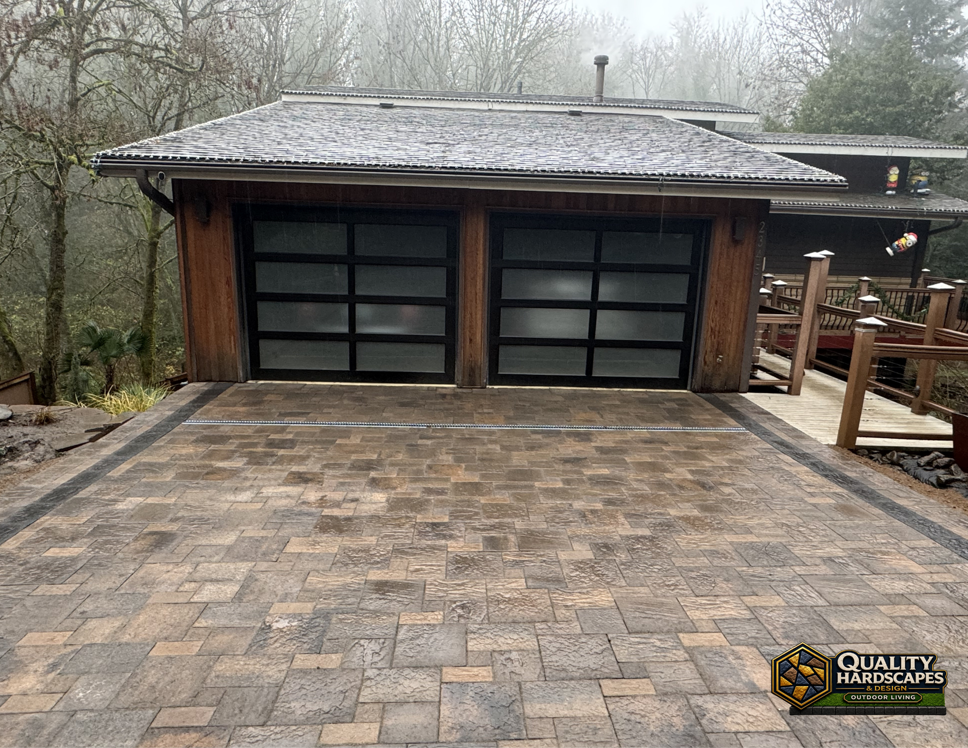 Paver driveway leads to a wood garage with frosted glass doors on a foggy, wooded property.