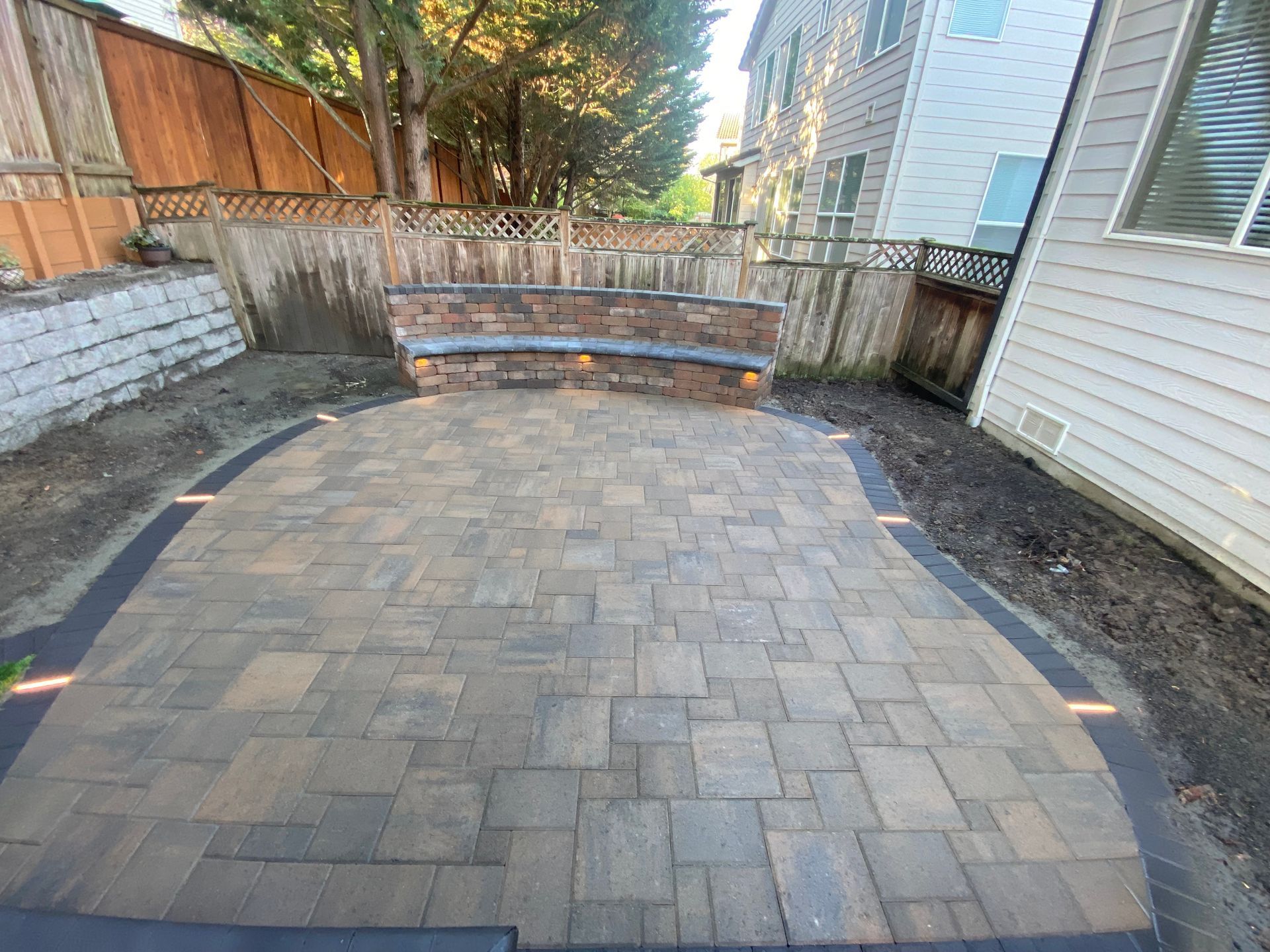 Paver patio with a built-in stone bench, bordered by a dark strip with lights, next to a wooden fence.