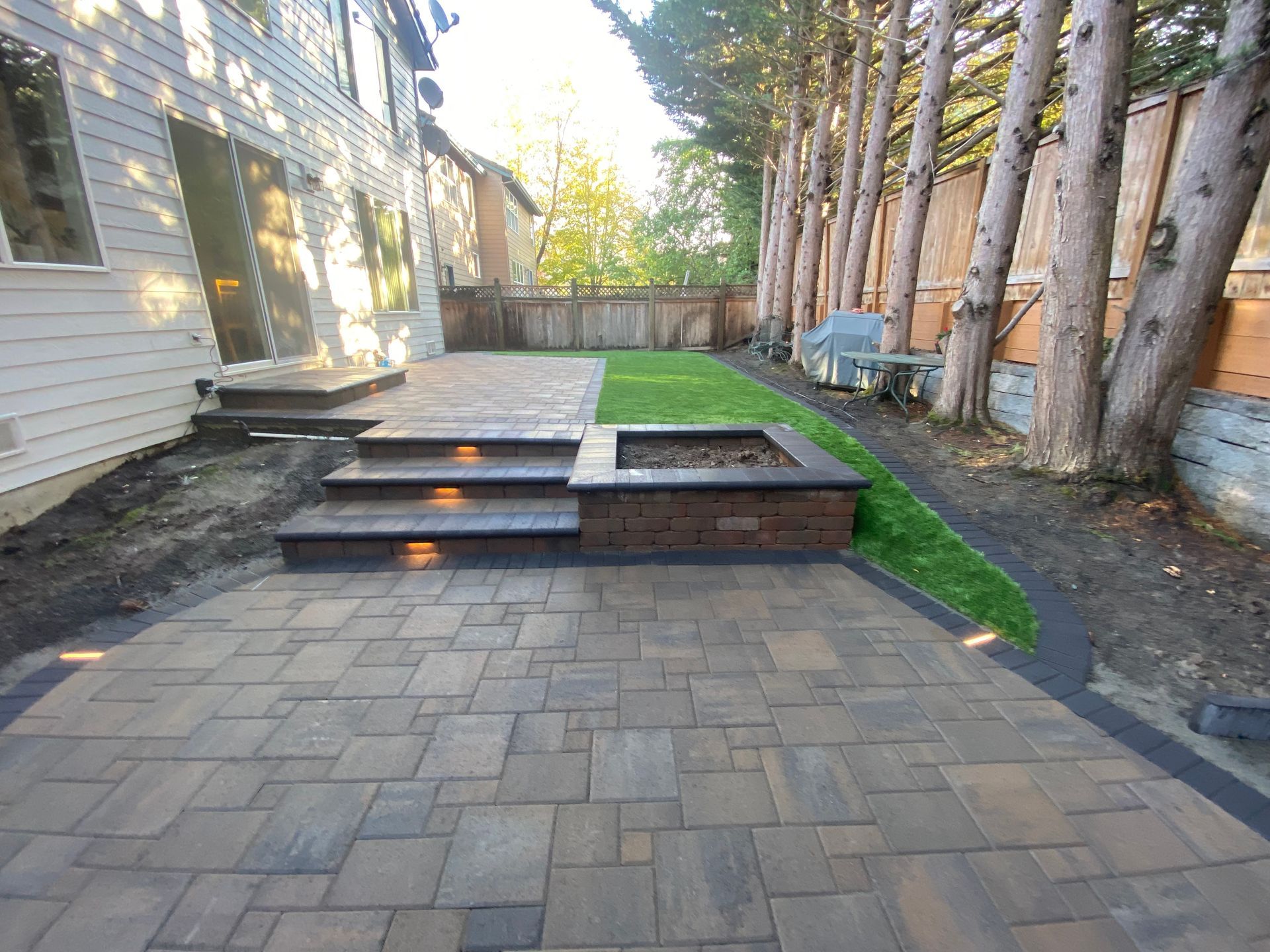 Backyard patio with brick pavers, steps, and a fire pit near a grassy area and trees.