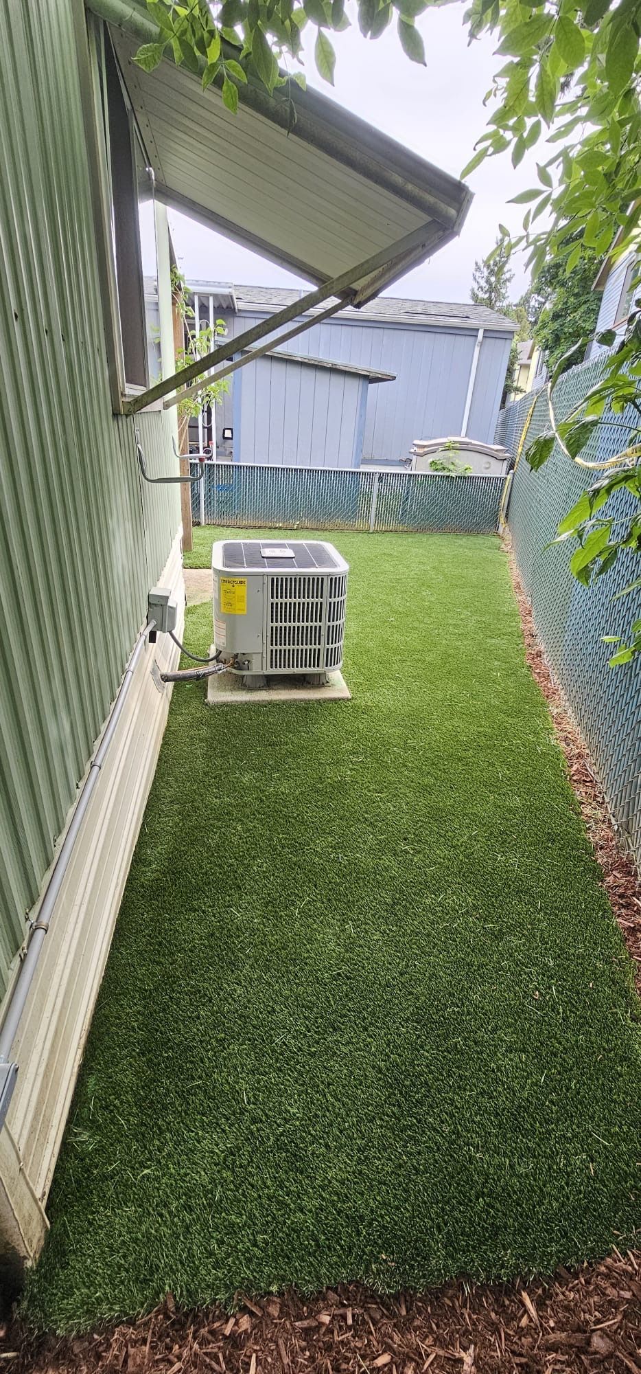 Narrow yard with green turf, AC unit, and chain-link fence. Awning on the side of a building.
