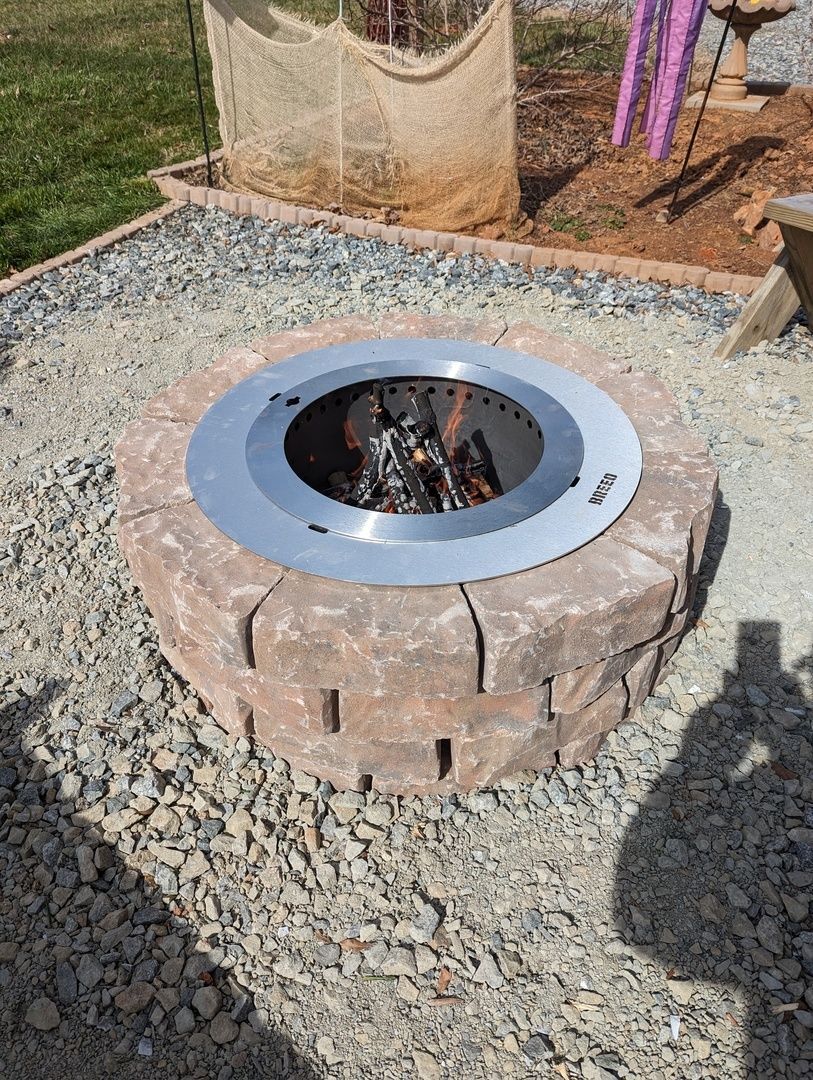 A round, brick fire pit with a metal ring cover containing burning wood and surrounded by gravel.