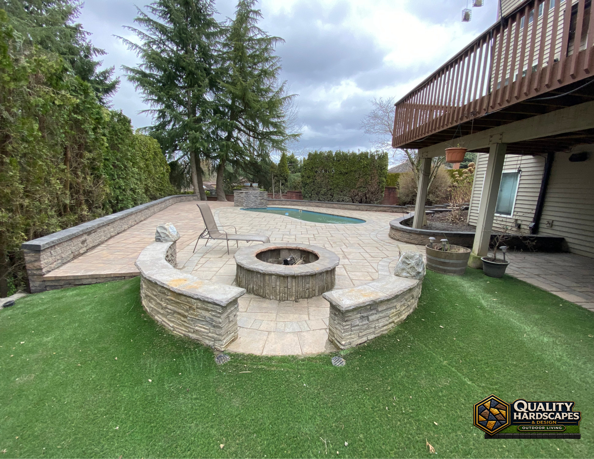 Stone fire pit seating area with curved benches on a patio beside a house and lawn