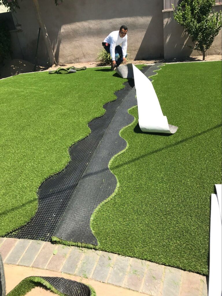 Man laying black mesh drainage on green artificial turf in backyard.