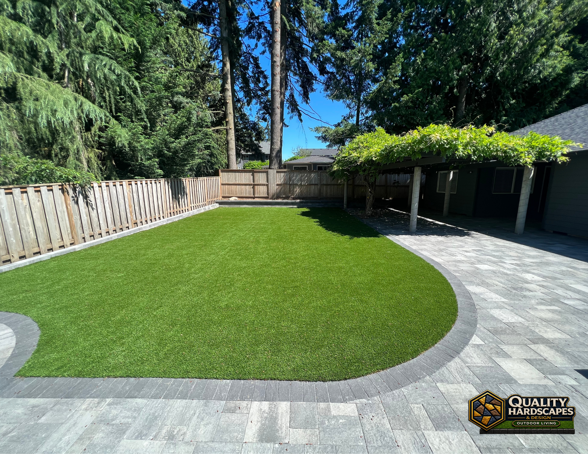 Curved green lawn bordered by gray pavers, wooden fence, and tall trees in a sunny backyard