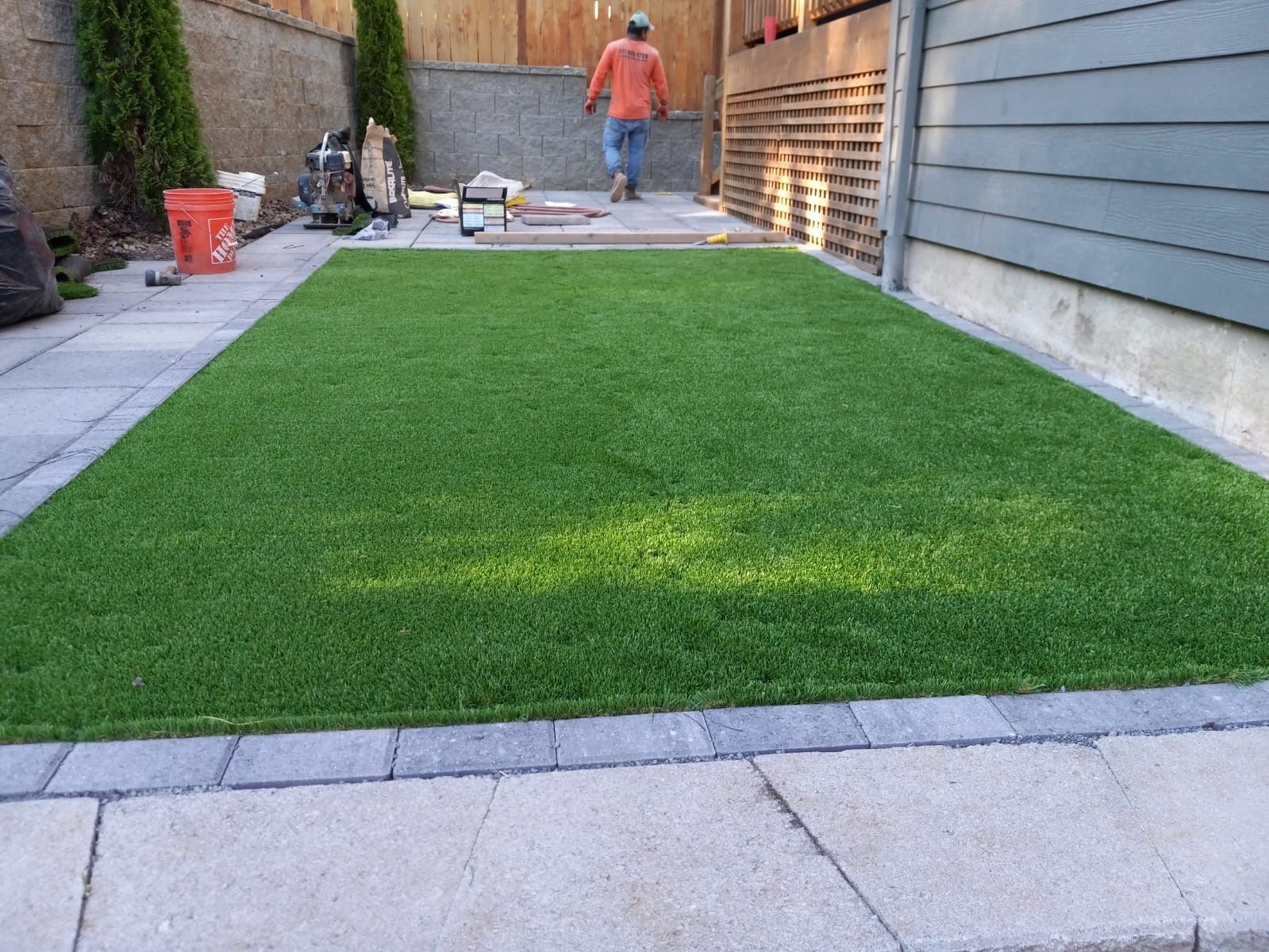 Artificial turf installed in a backyard, bordered by pavers. A person is walking in the background.
