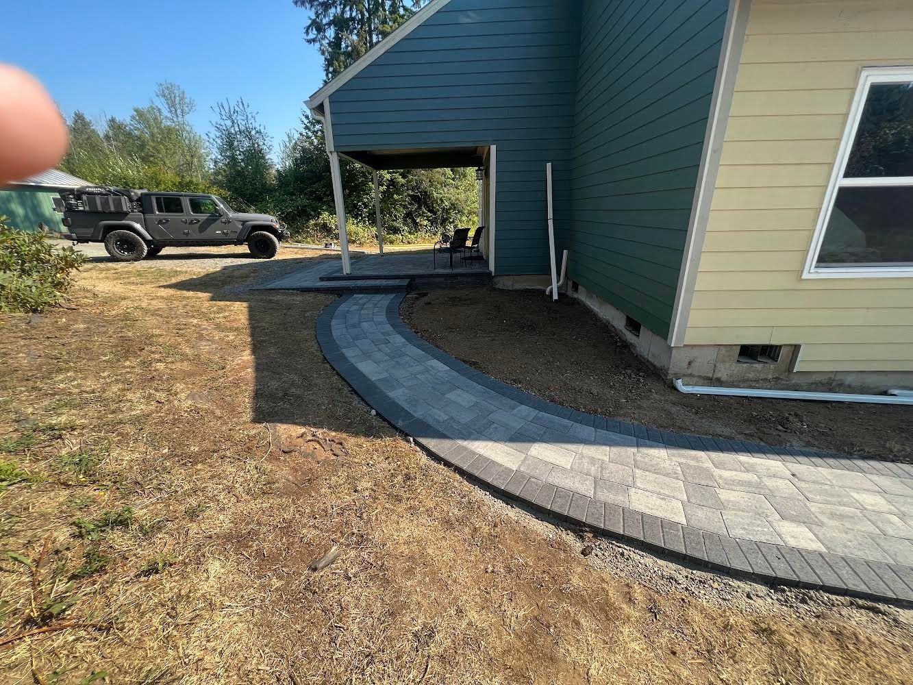 Brick pathway curves alongside a house with green and yellow siding; a vehicle is in the background.